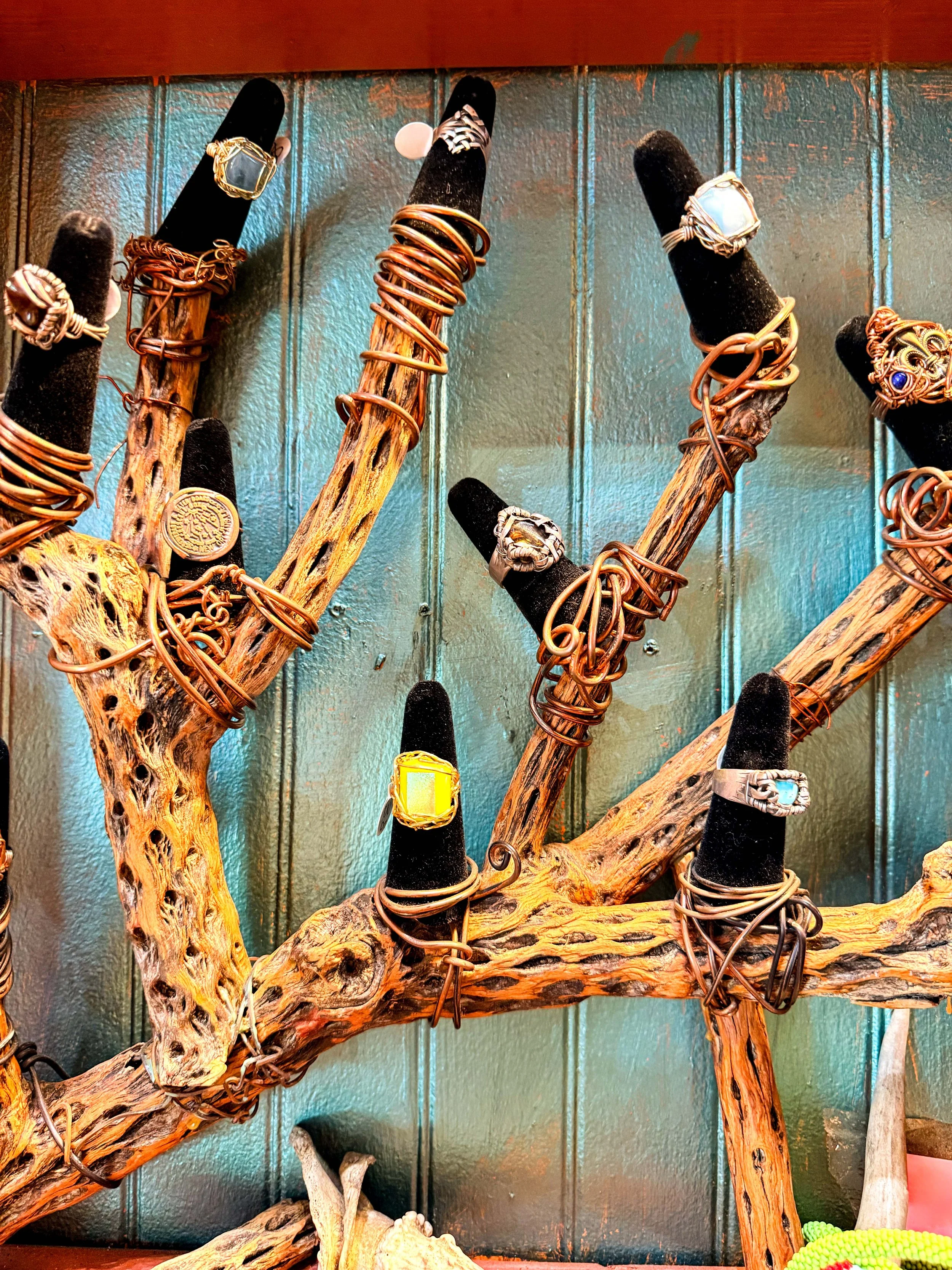 Display of jewelry rings on black velvet finger mannequins mounted on a rustic wooden stand with a blue-green background.