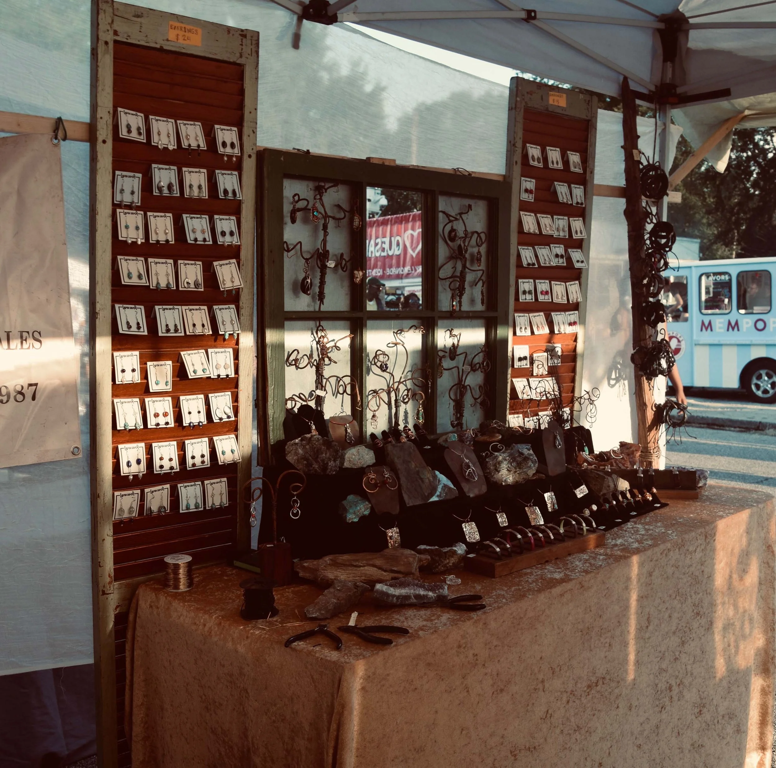 Jewelry display at an outdoor market stall, featuring handmade earrings, rings, necklaces, and bracelets arranged on boards and rocks, with pliers and jewelry-making tools on the table.