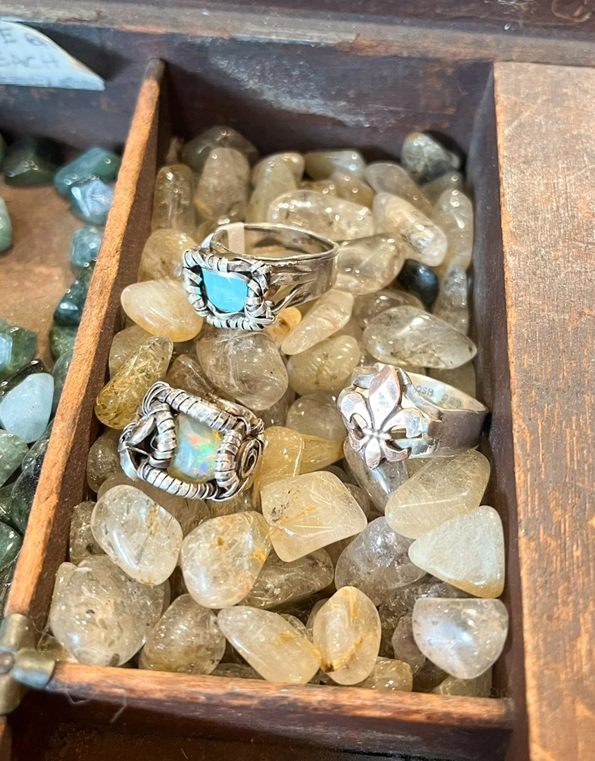 A wooden box containing polished yellowish stones with three silver rings on top, one with a turquoise stone, one with an opal, and one with a floral design.
