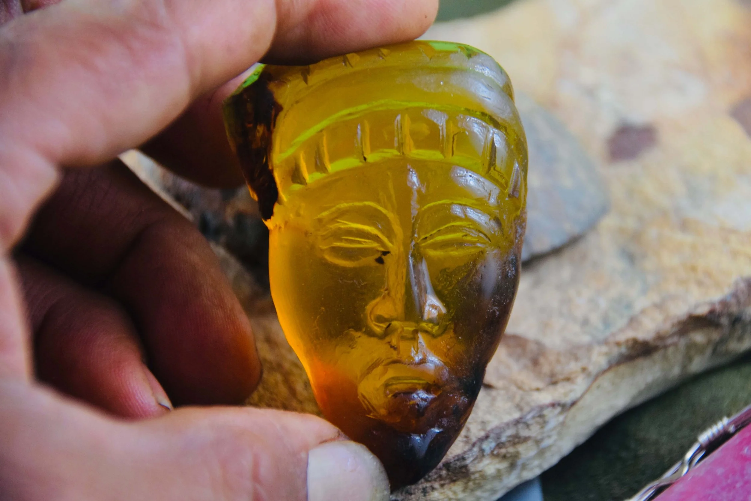 A close-up of a hand holding a carved piece of amber in the shape of a human face, with detailed facial features and a headdress, resting on a stone surface.