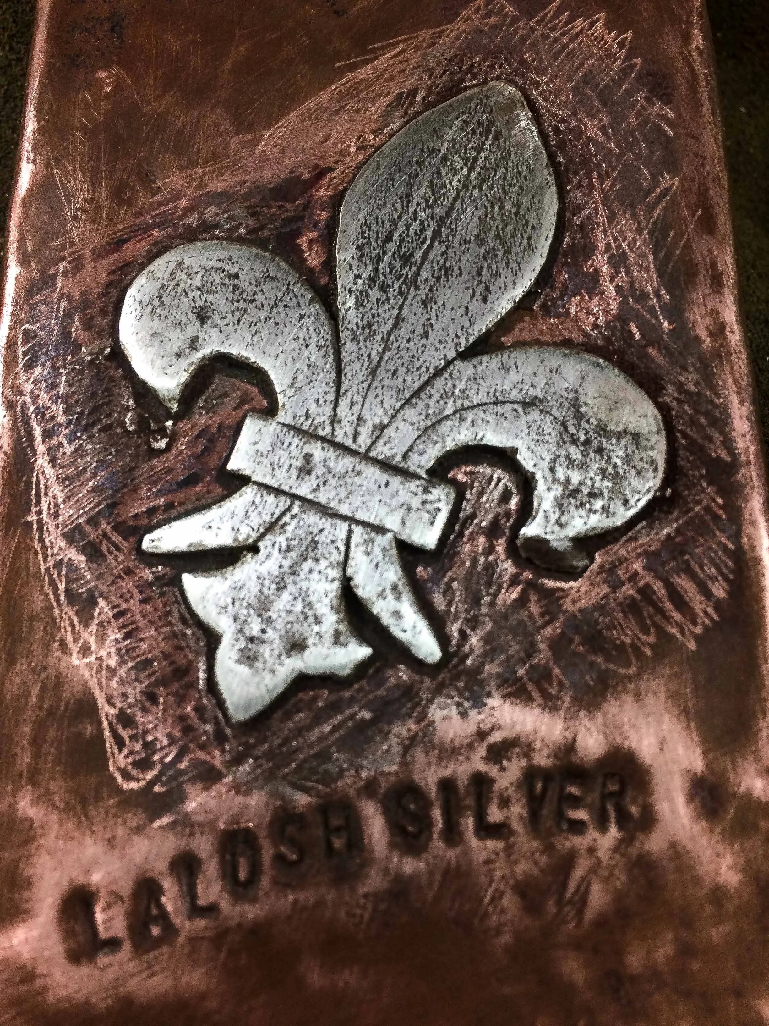 A metallic emblem of a fleur-de-lis symbol with the words 'LALOS SILVER' engraved below, set against a textured background.