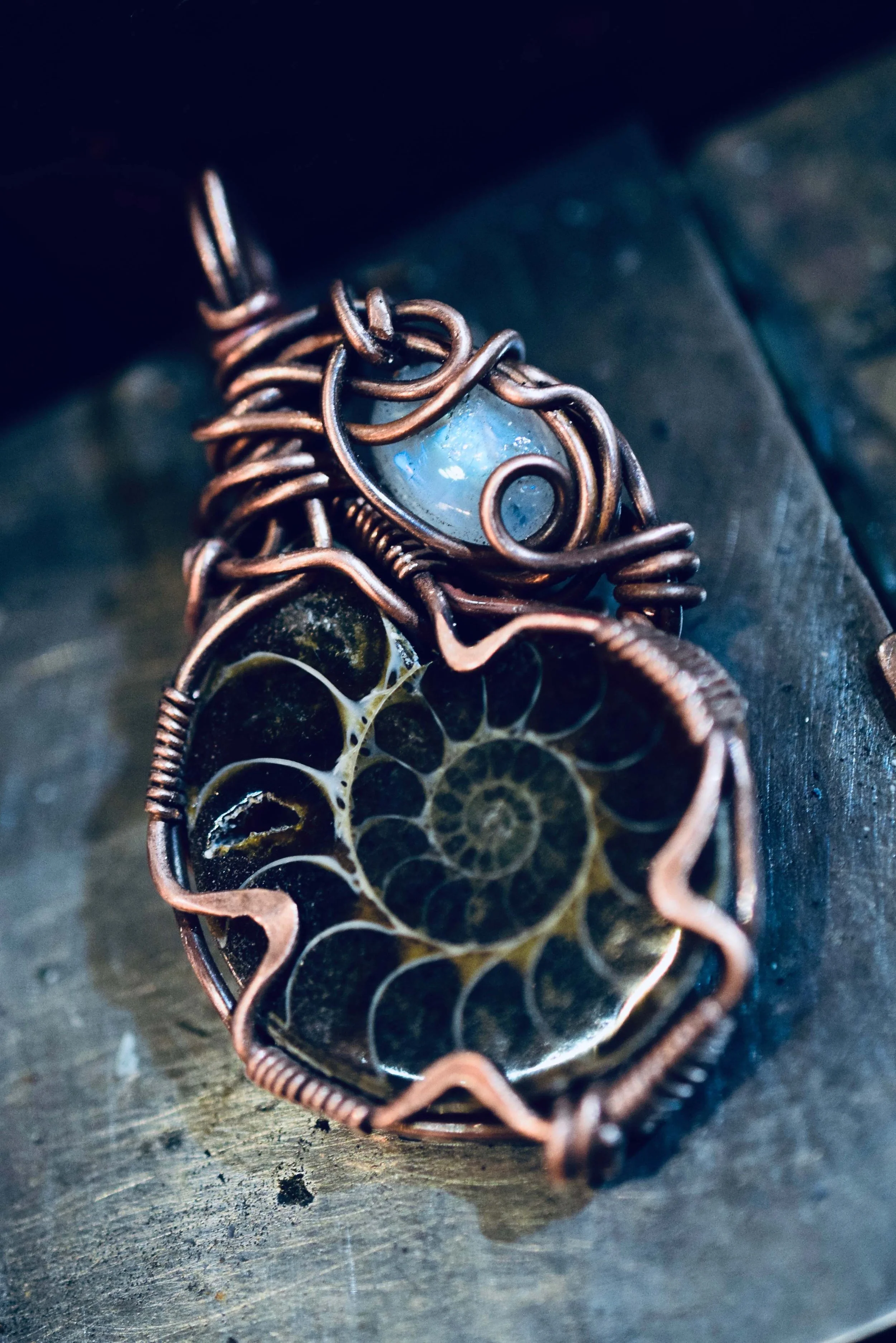 Close-up of a wire-wrapped pendant with a spiral design inside. The pendant has an oval, light blue stone at the top, surrounded by copper wire, and a black and white patterned spiral in the center.