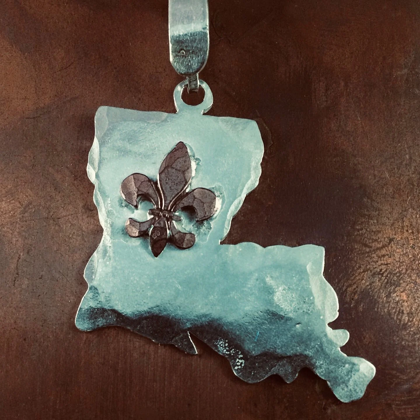 Metal keychain in the shape of the state of Louisiana with a black fleur-de-lis symbol in the center, lying on a wooden surface.
