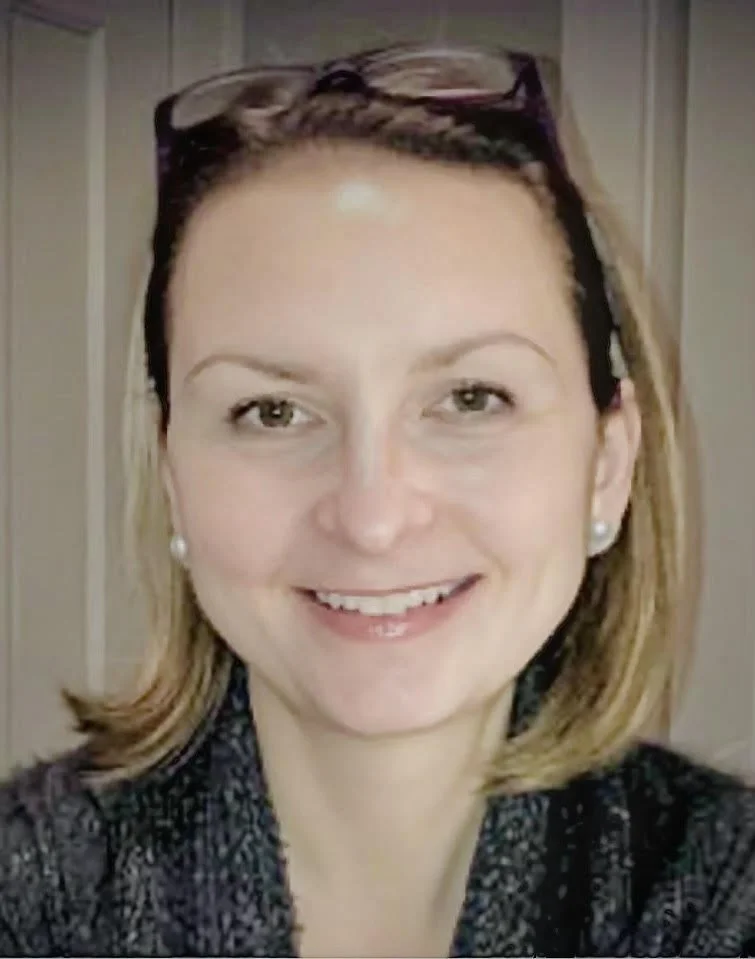 A woman with shoulder-length blonde hair, light-colored eyes, wearing pearl earrings, sunglasses on her head, and a dark patterned top, smiling at the camera.
