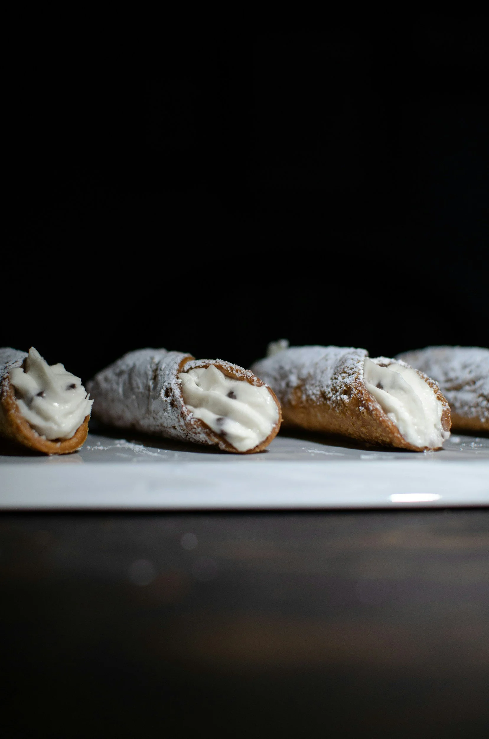 Three cannoli filled with cream on a white plate, dusted with powdered sugar, against a black background.