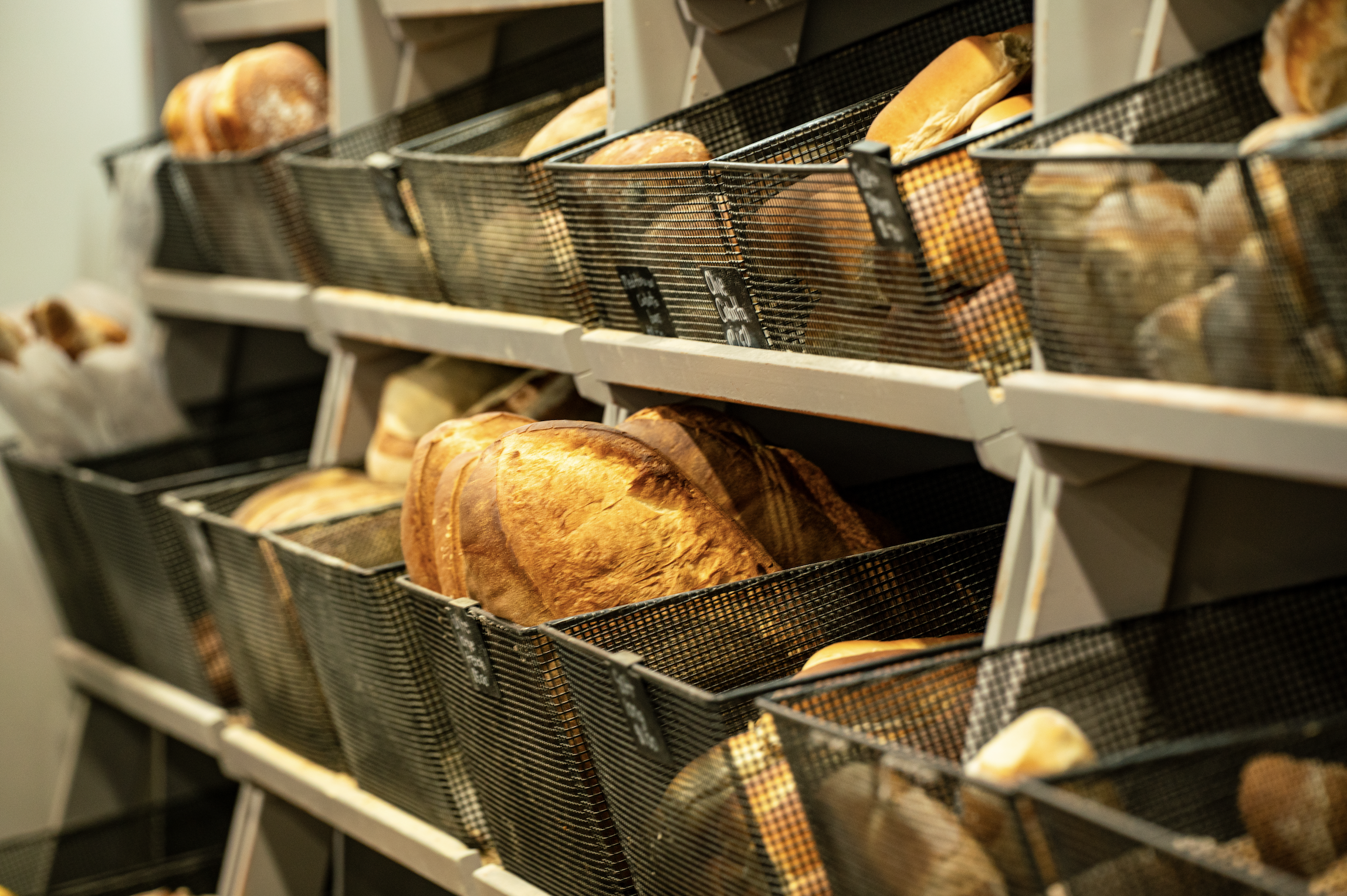 Display of various loaves of bread stored in black wire baskets on white shelves, in a bakery or grocery store setting.