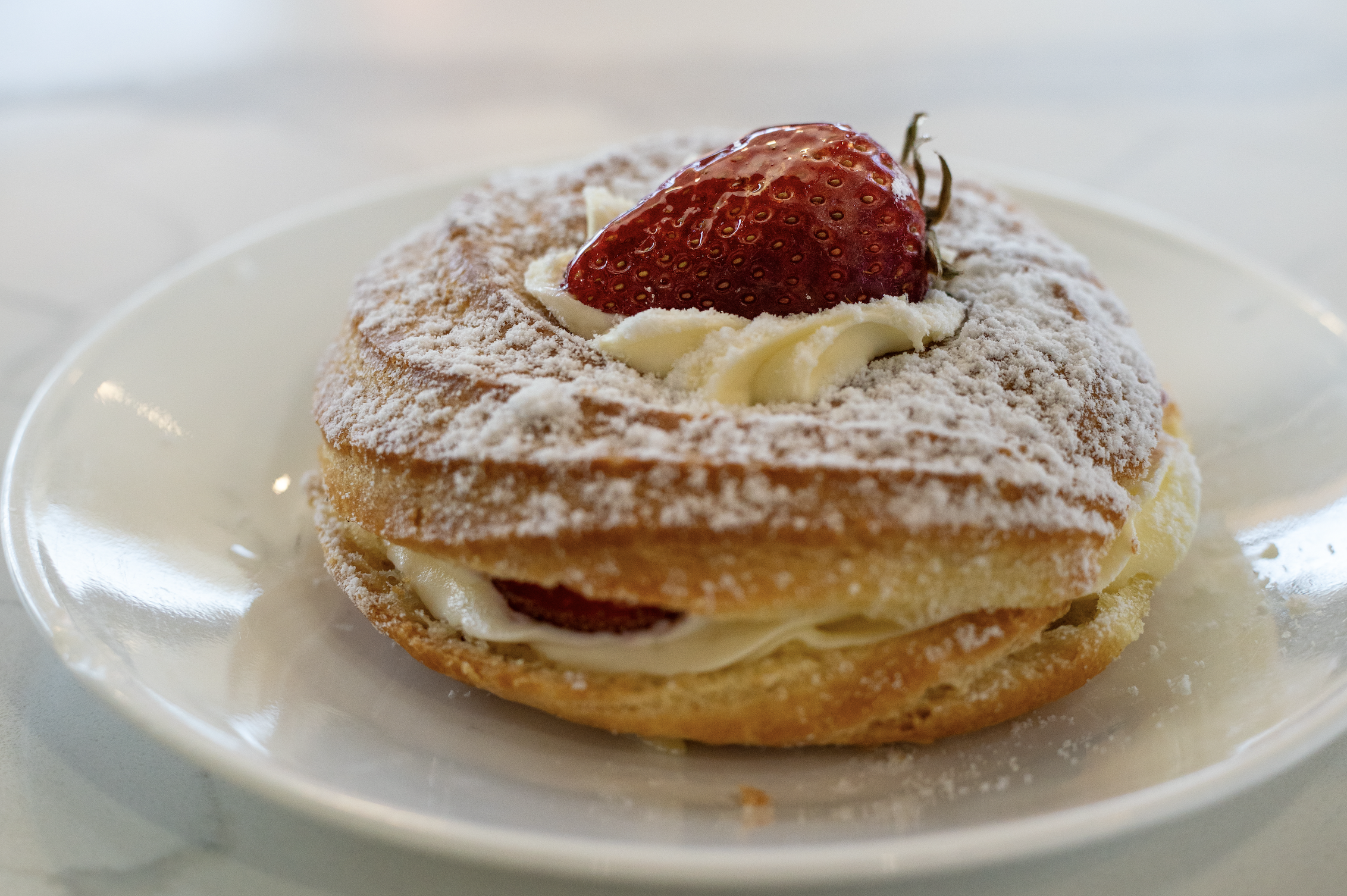 Close-up of a cream-filled pastry topped with powdered sugar, a sliced strawberry, and white chocolate shavings on a white plate.