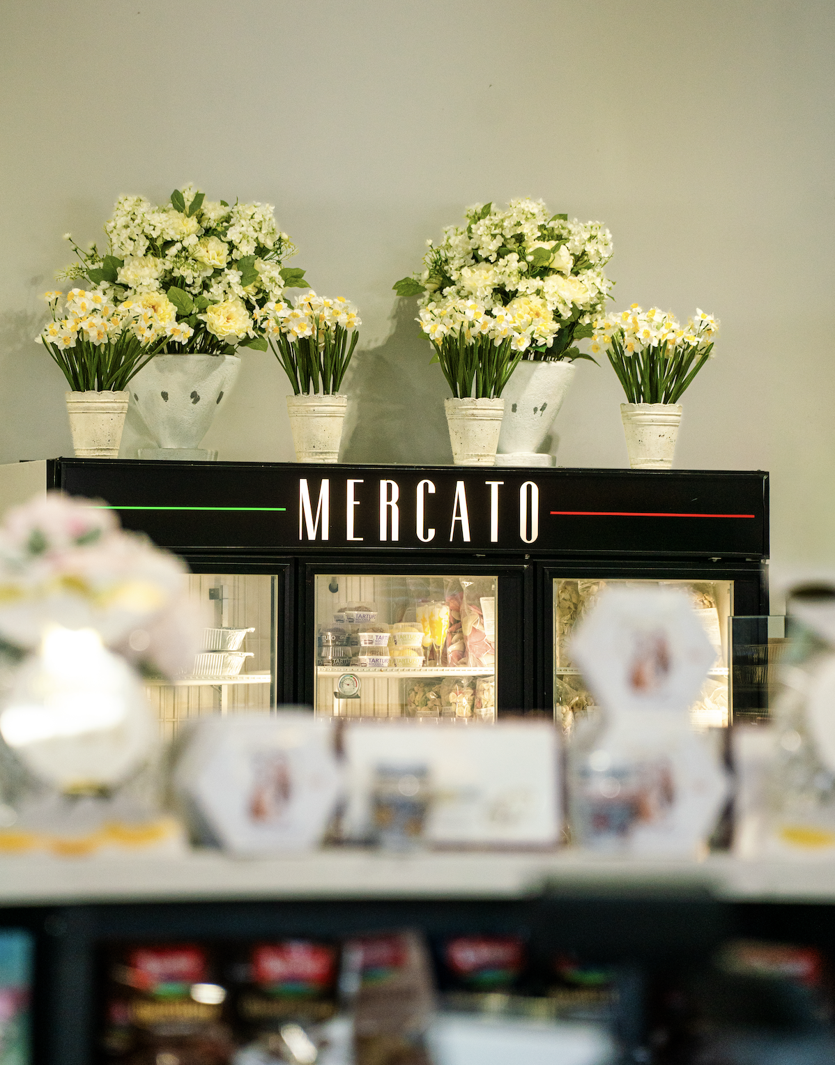 Fresh flower arrangements with white and yellow blooms on display above a refrigerated counter labeled 'Mercato' in an indoor market or grocery store.