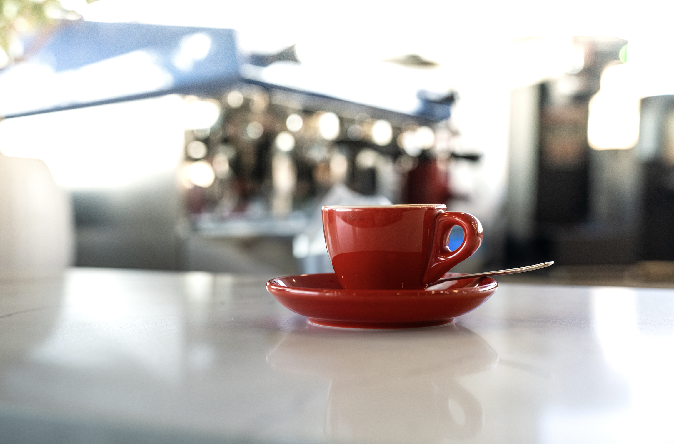A bright red coffee cup with a matching saucer on a white surface, with a blurred background of a coffee shop or cafe.