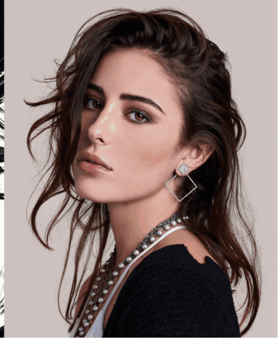A woman with dark wavy hair, wearing earrings and layered necklaces, wearing a black top, looking at the camera.