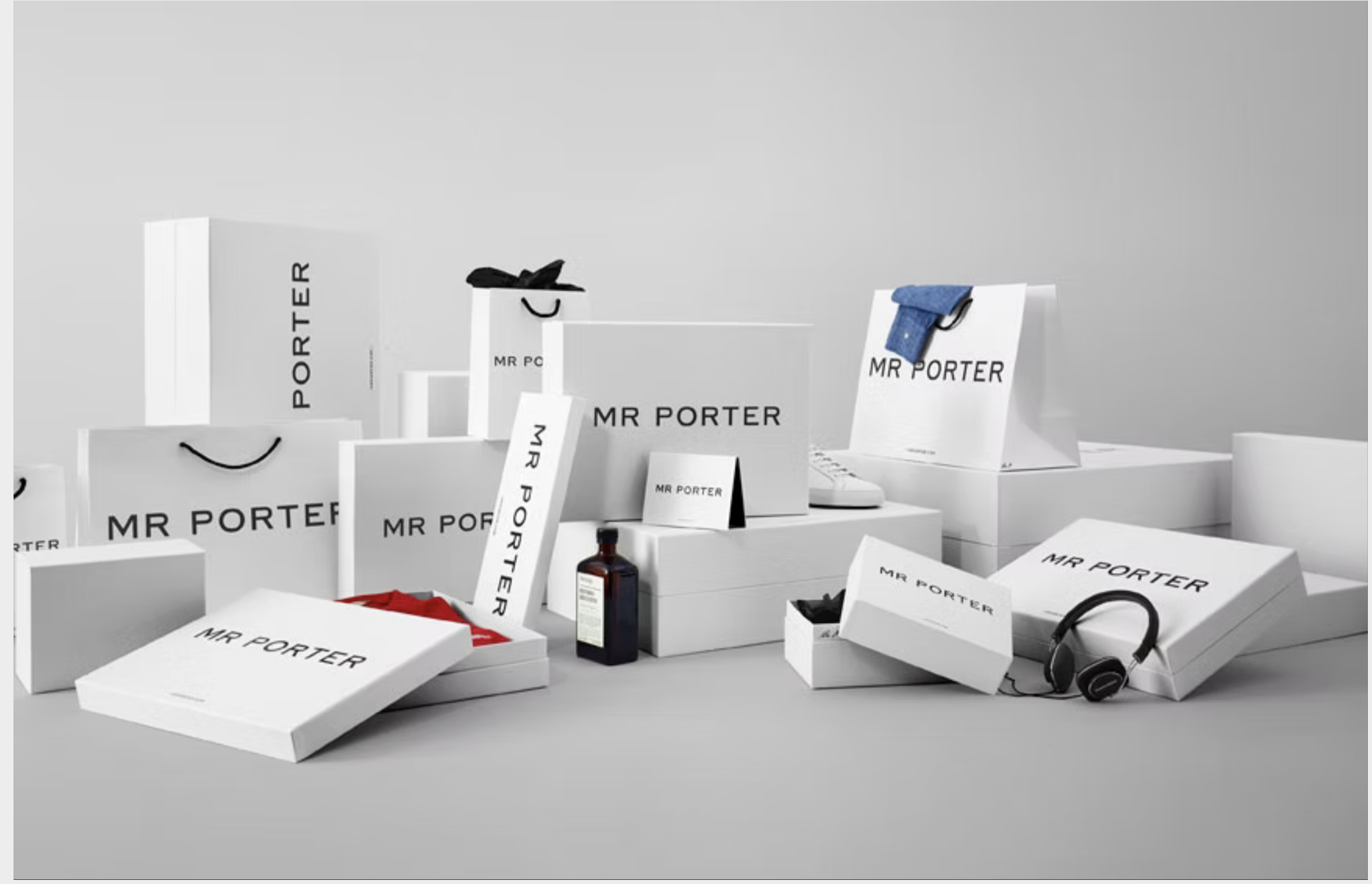Various white shopping bags, boxes, and packaging with 'Mr Porter' branding, some with black handles, a pair of black headphones, and a perfume bottle on a gray surface against a plain gray background.