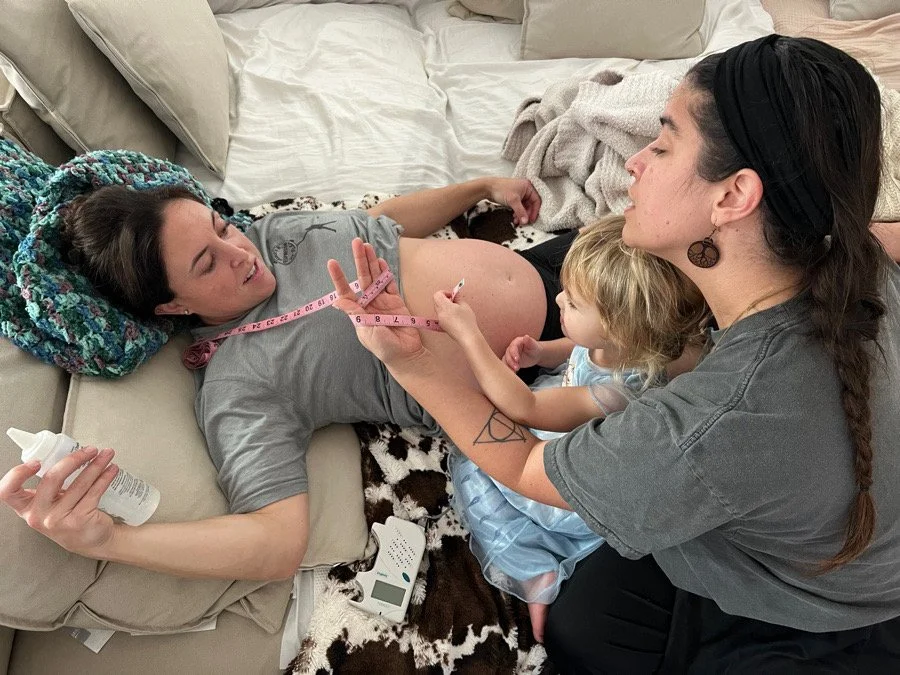 Pregnant woman lying on a couch with her hand on her belly, receiving a blood pressure test from a woman with a child sitting beside her.