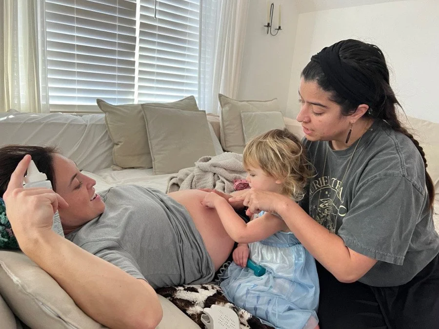 A woman lying on a couch with a pregnant belly, a young girl touching her belly, and a woman kneeling beside them gently touching the girl's arm, in a cozy living room with beige pillows and white walls.