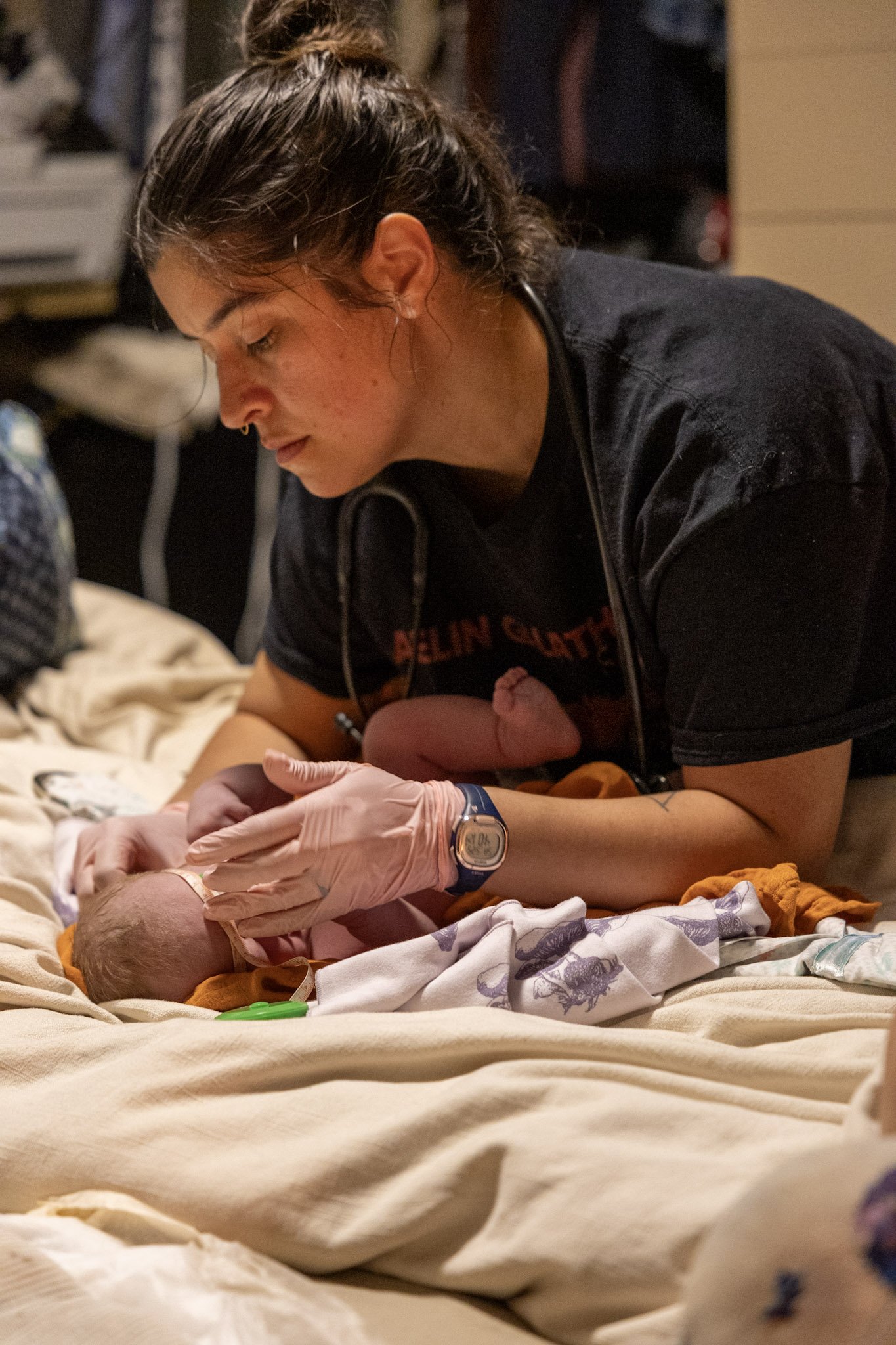 A woman in a black t-shirt with a stethoscope around her neck is holding a newborn baby wrapped in a blanket, on a bed in a hospital or medical setting.
