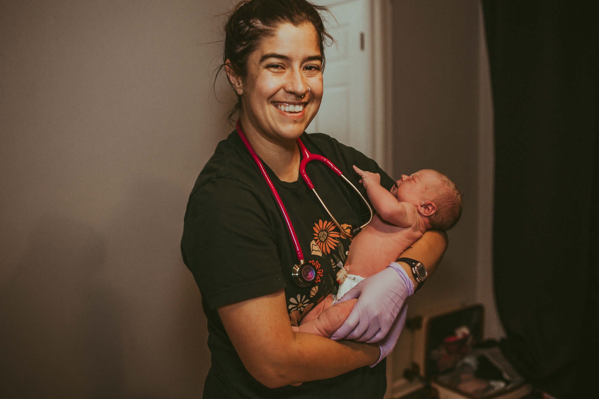 Dani Wright Midwife smiling with a stethoscope around her neck holding a newborn baby in her arms.