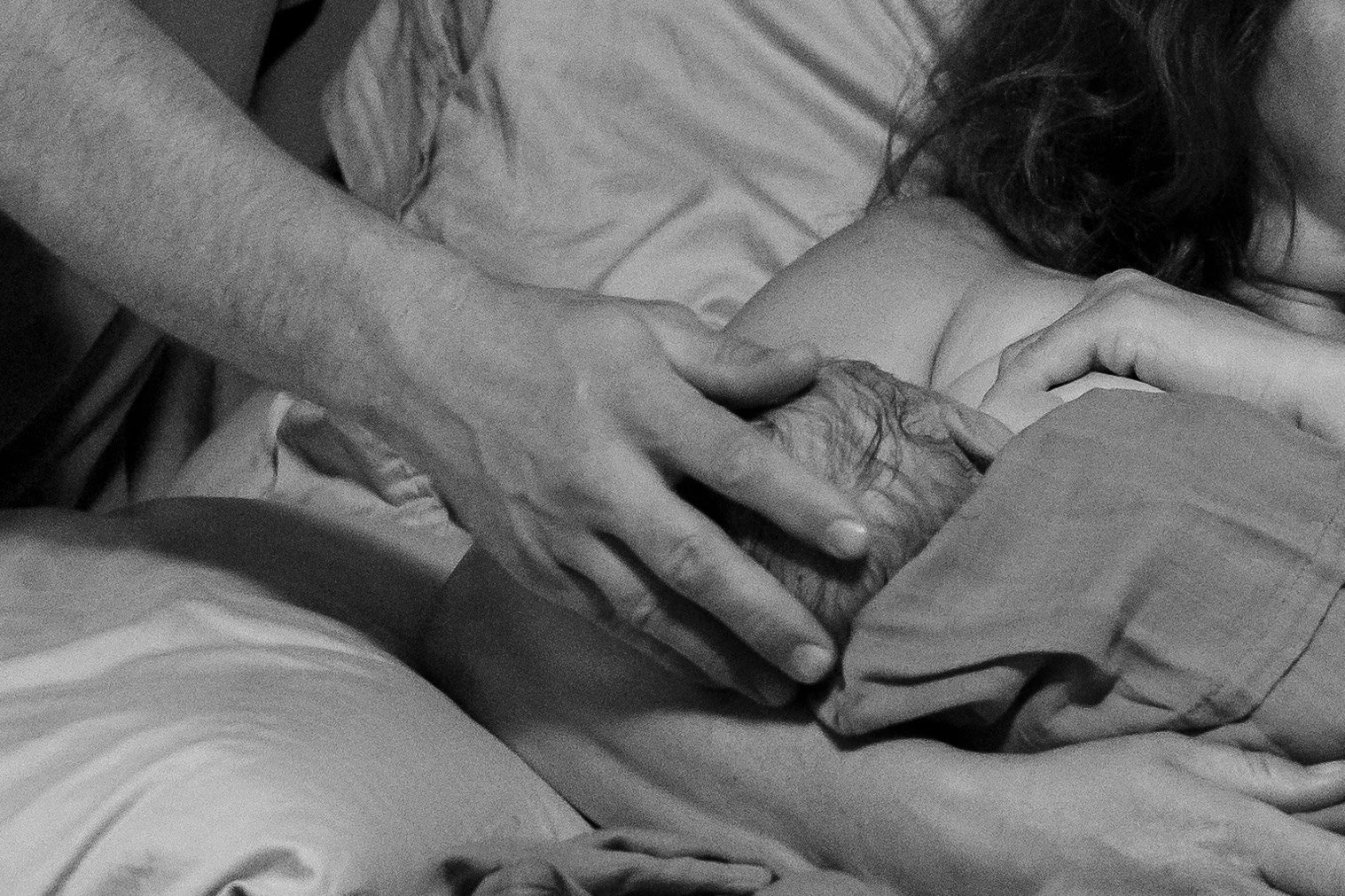 A black and white photograph showing two pairs of hands, one appearing older with wrinkles, gently holding the arm of a young woman with long hair, lying on a bed.