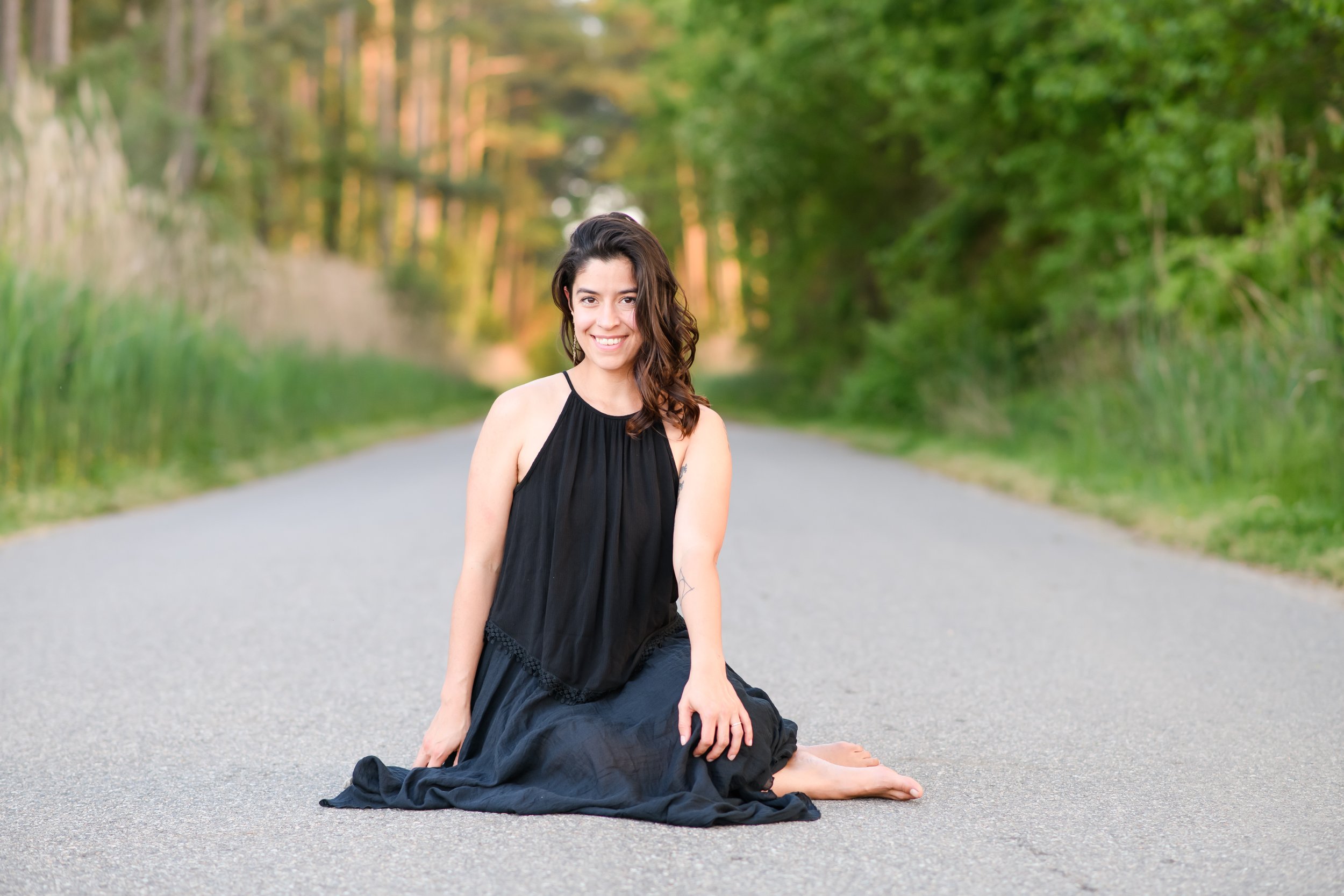 Dani Wright midwife in a black dress sitting on a paved road in a forested area, smiling at the camera with trees and greenery in the background.