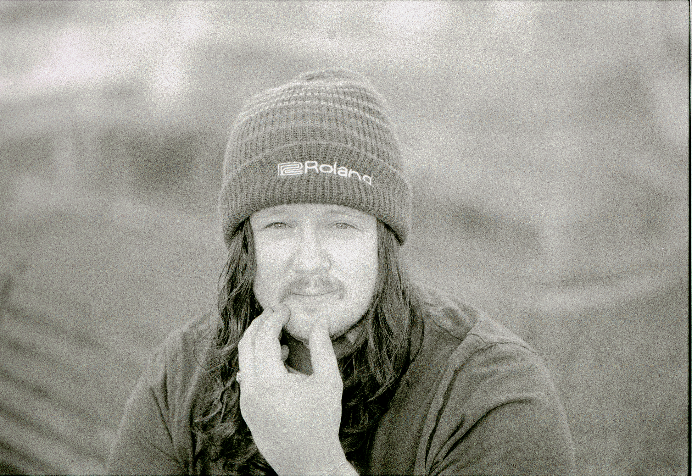 A man with long hair and a beard, wearing a knit beanie hat and a jacket, sitting outdoors with a cloudy sky in the background. Shot on Kodak P3200 TMax 35mm film stock.