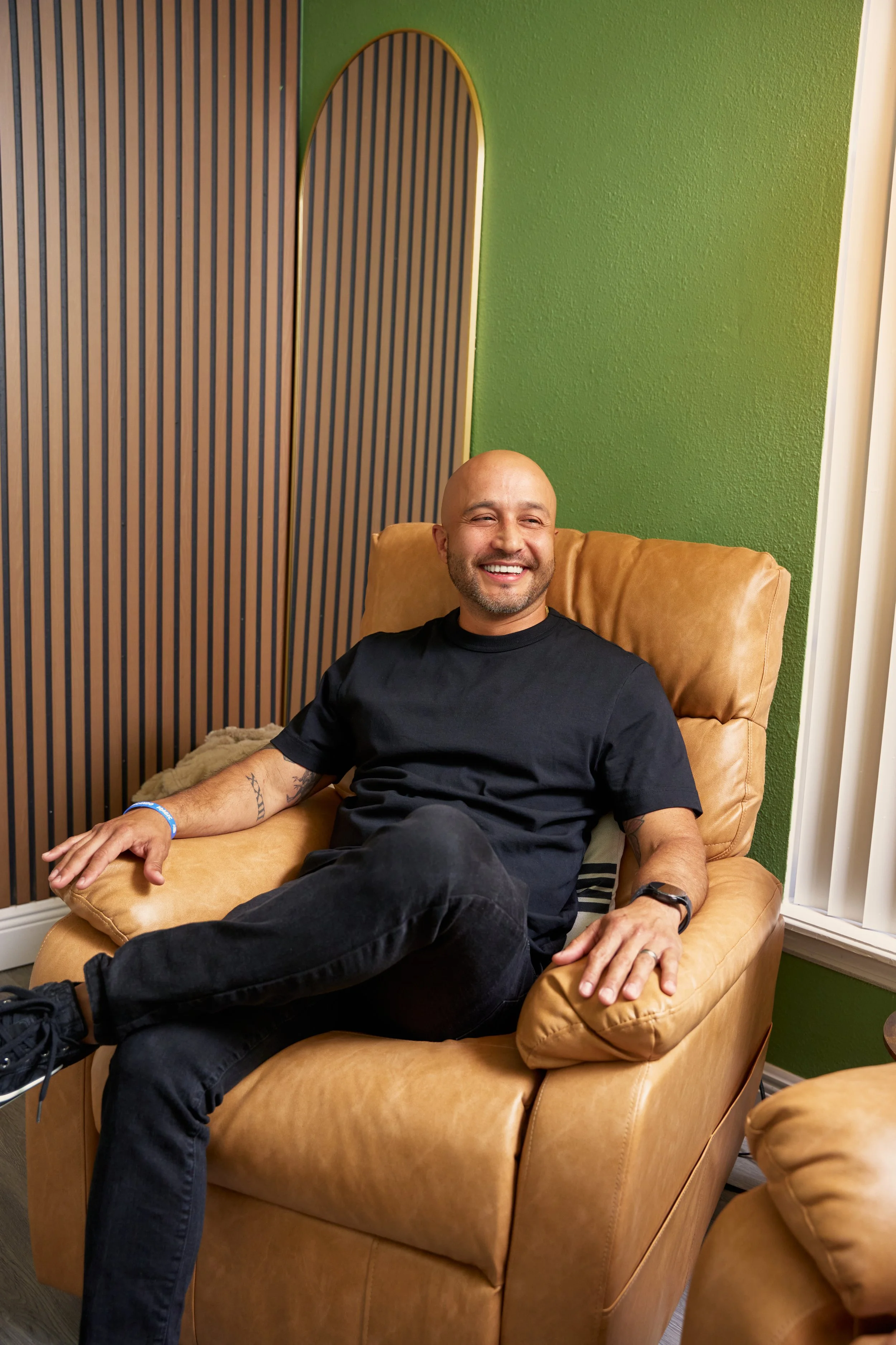 A bald man with a beard, wearing a black t-shirt and black jeans, sitting on a tan leather chair in a room with green walls and unique wooden paneling, smiling while looking to his left.