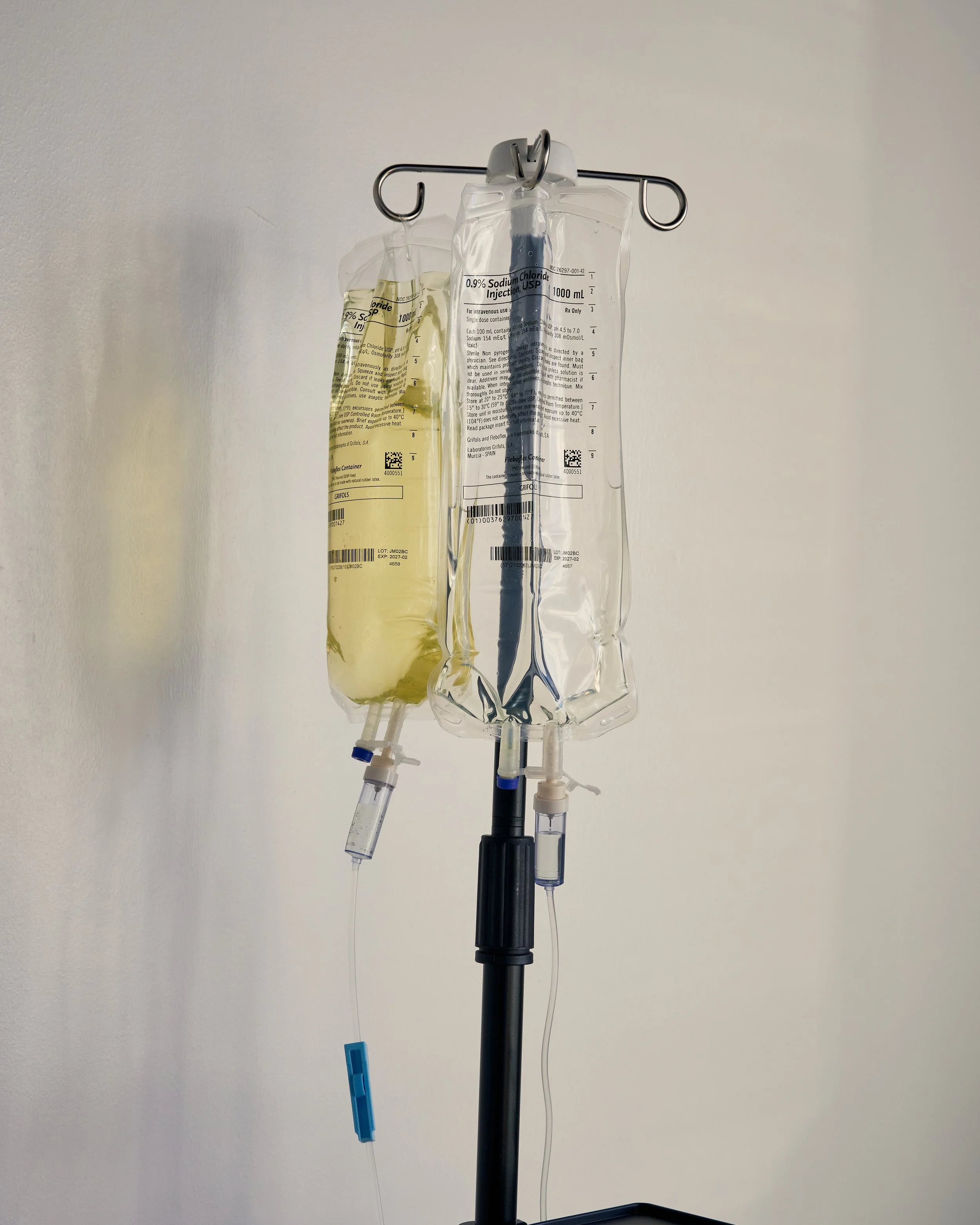 IV drip set with yellow and clear fluids hanging from a pole against a plain wall.