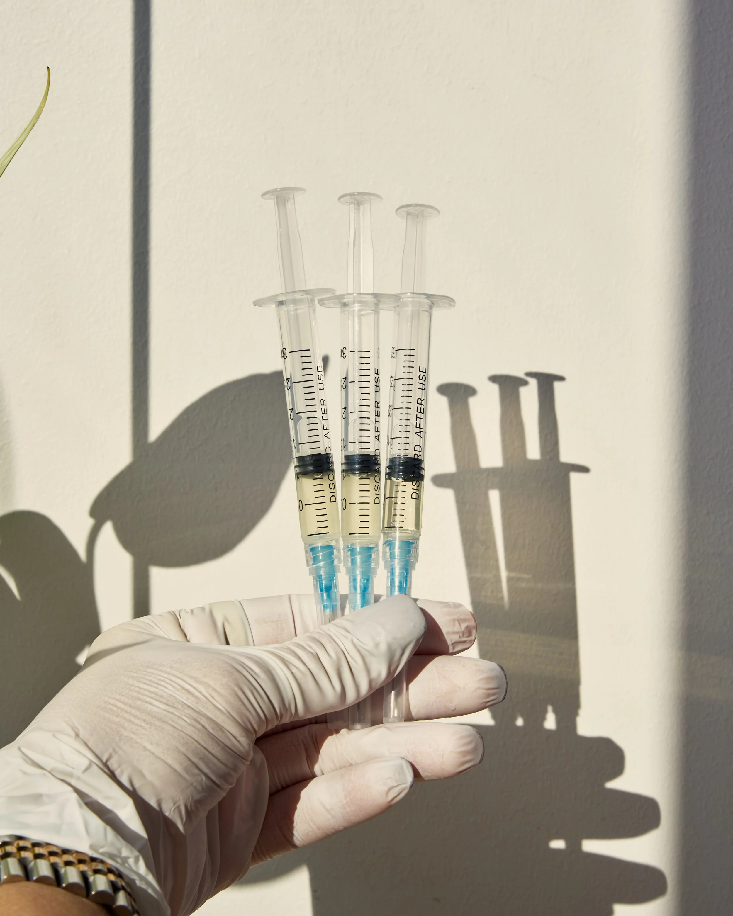 A hand in a white glove holding three syringes filled with a yellowish liquid, with shadows cast on a white wall.