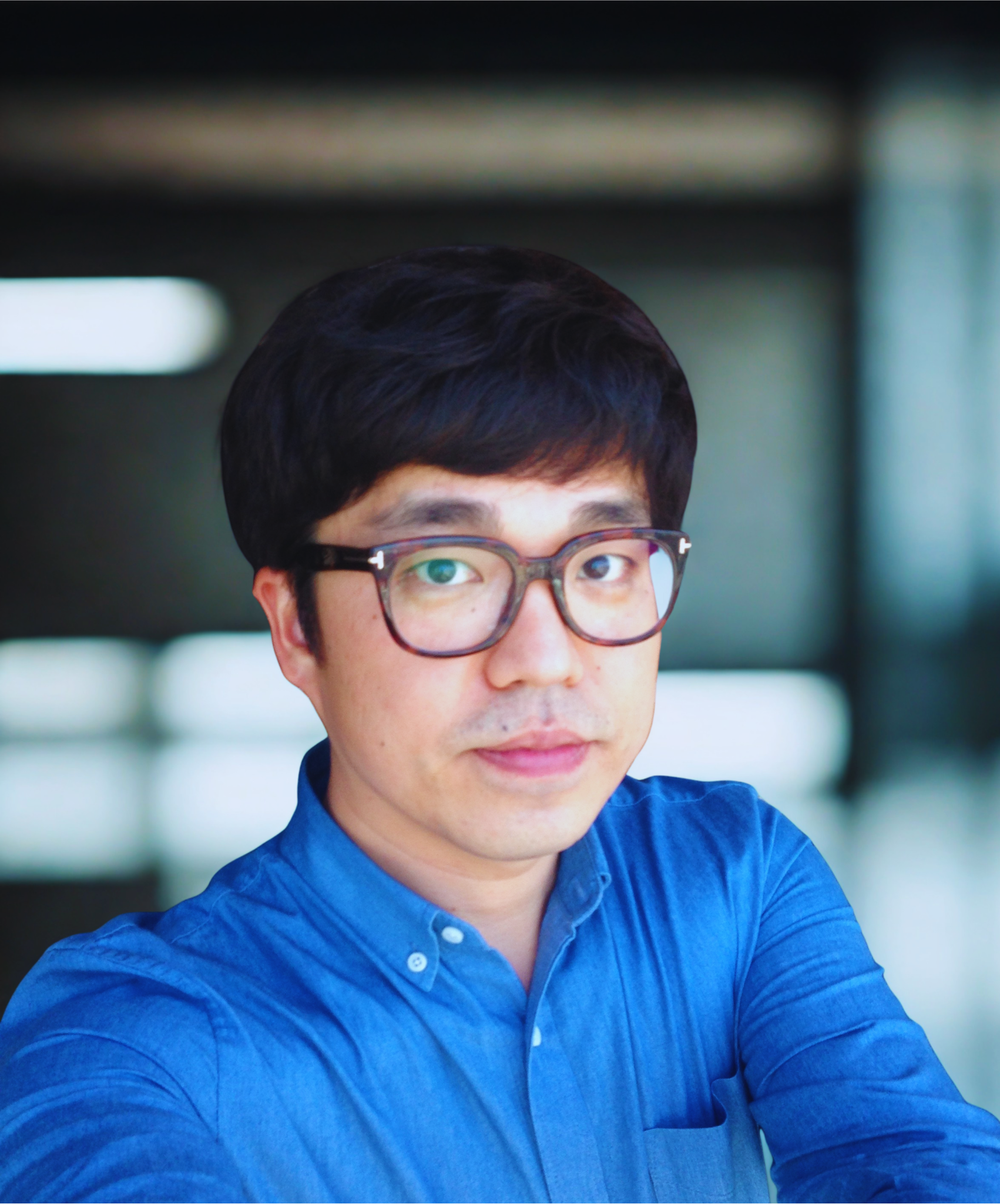 A portrait of a man wearing glasses and a blue button-down shirt, with dark hair, looking into the camera, in an indoor setting with blurred background.