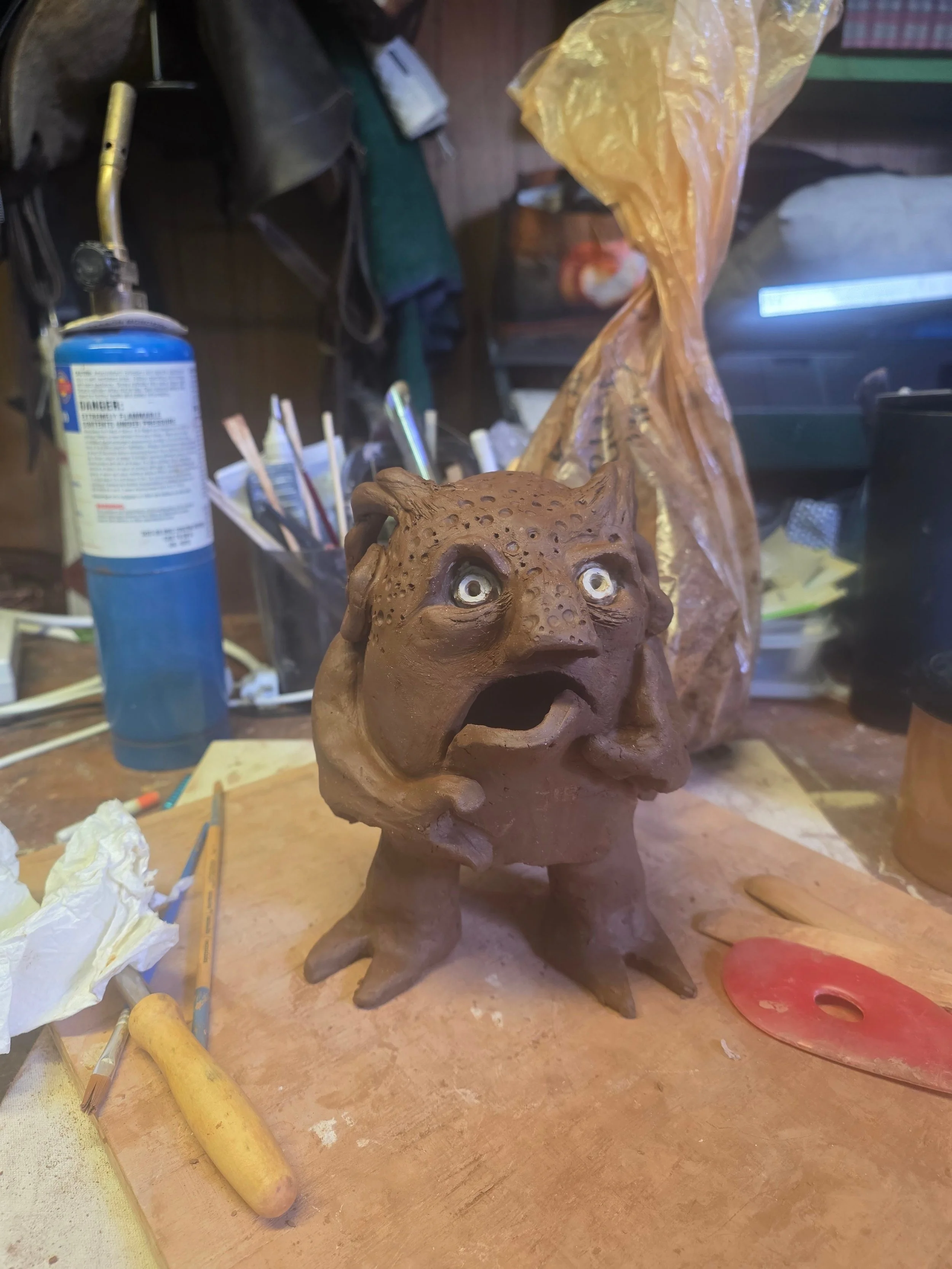 Unfinished ceramic sculpture of a creature with a human-like body and a head resembling a fish or amphibian, with large, expressive eyes and an open mouth. Speckled Buff clay body.