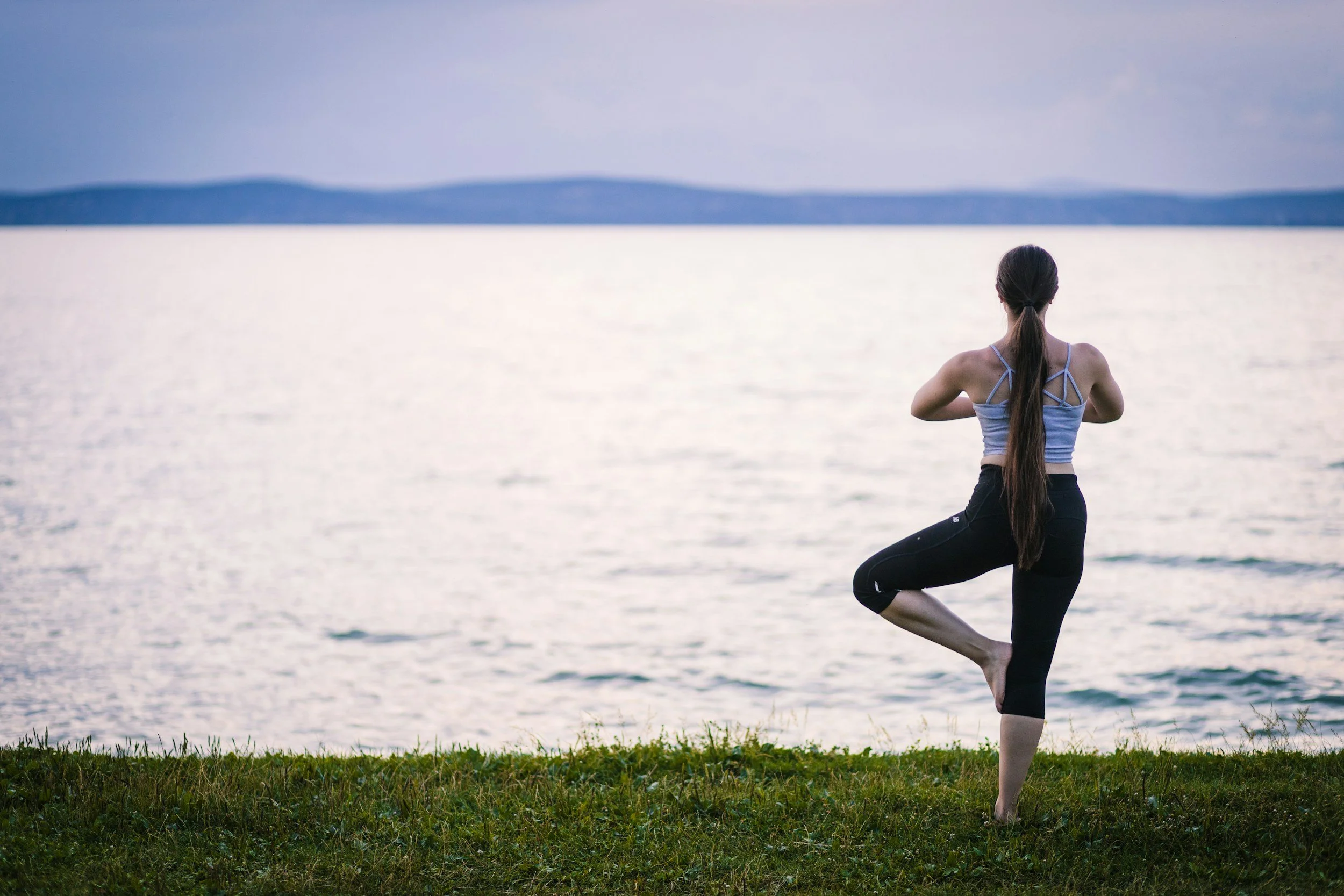 A woman practicing yoga outdoors on grass near a body of water, performing the tree pose with one foot resting on her inner thigh and arms in prayer position, with a distant view of mountains across the water.