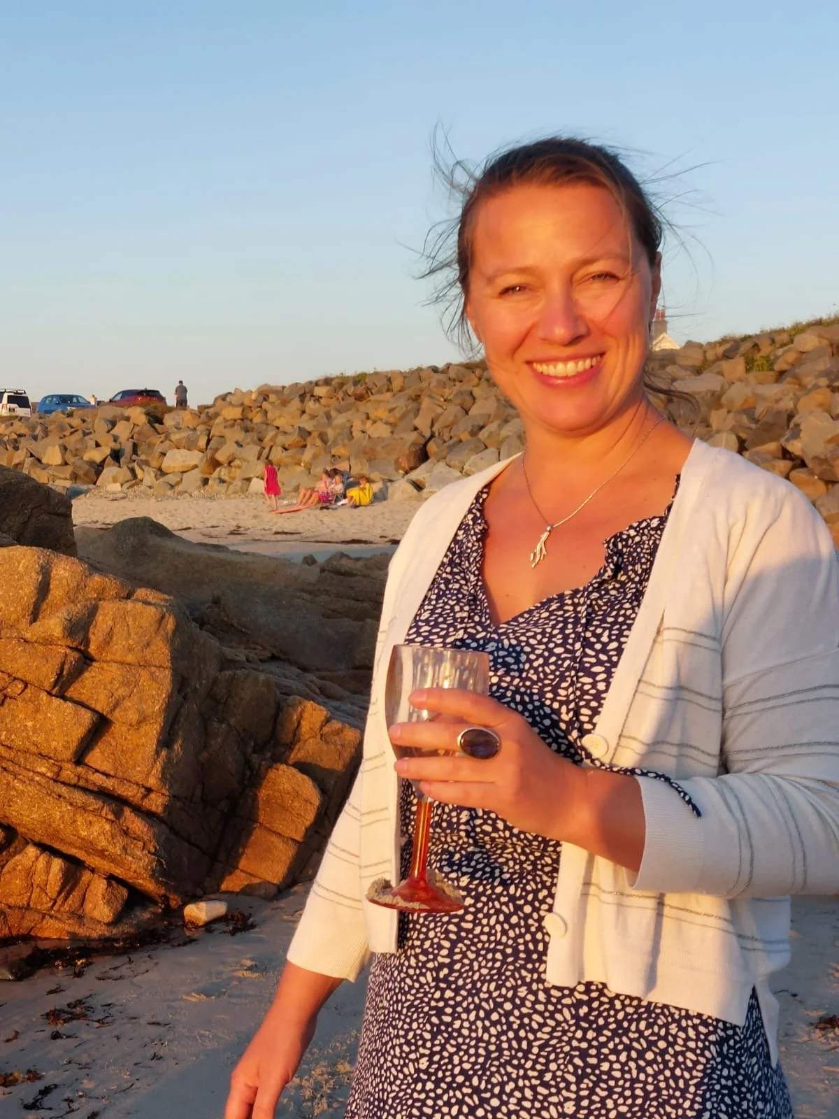 Woman smiling on the beach holding a glass of wine during sunset with rocky shoreline and cars in the background.
