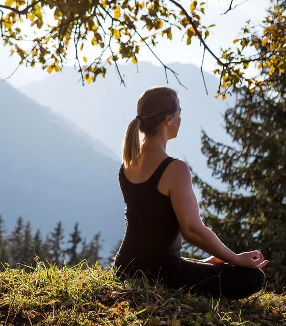 Woman meditating outdoors on grass, sitting cross-legged with closed eyes, trees and mountains in the background, autumn leaves on tree branches overhead.