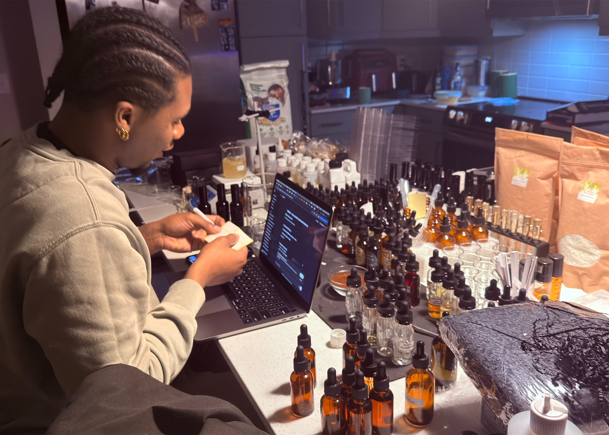Person with braided hair and gold hoop earrings preparing essential oils and ingredients for a product in a lab or workshop setting, surrounded by bottles, packaging, and equipment.