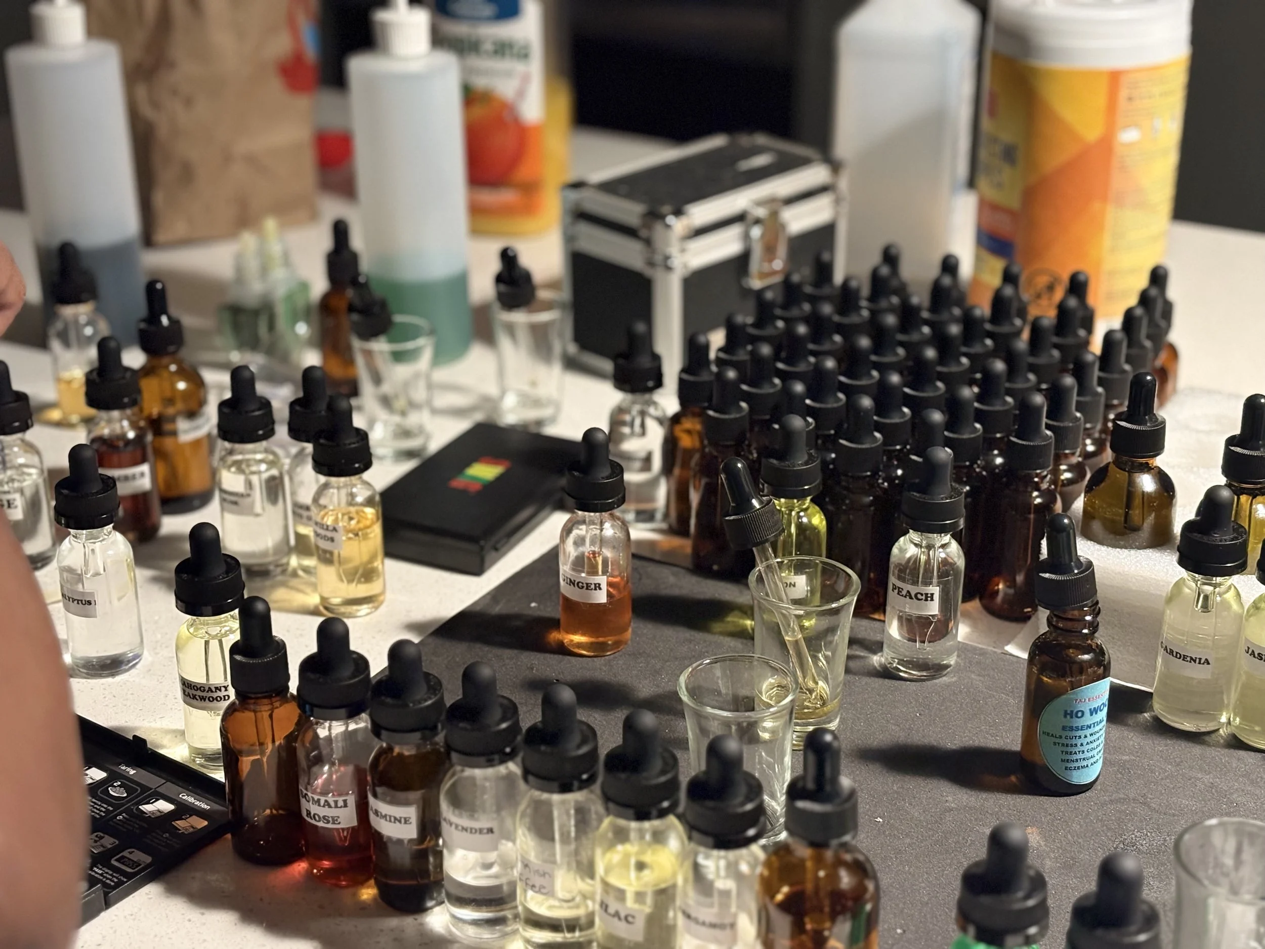 A collection of essential oils and aroma samples in small glass bottles with dropper caps, some bottles labeled with scent names like ginger, peach, and rose, set on a table with some measuring tools and containers in the background.