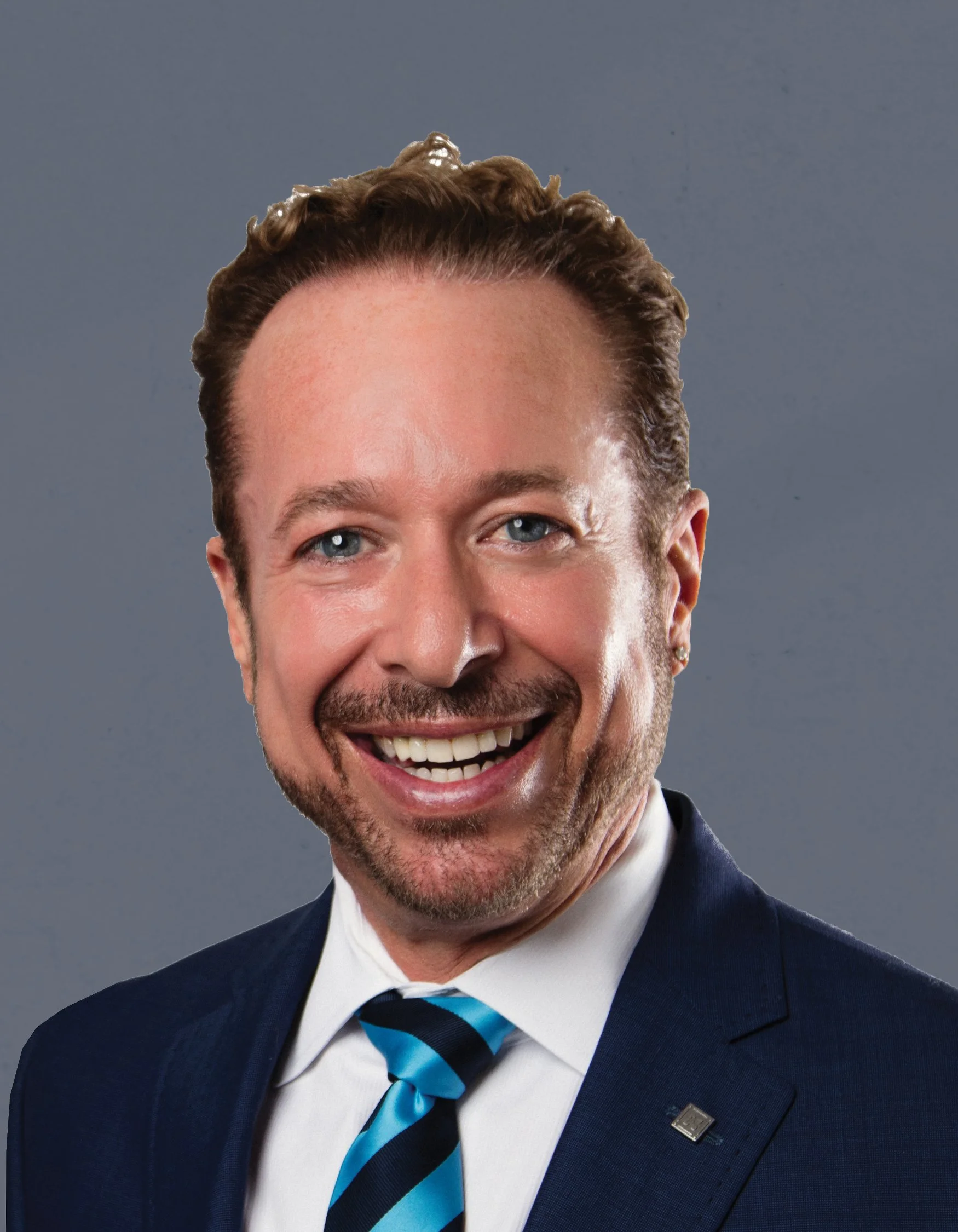 A middle-aged man with short wavy brown hair, blue eyes, and a beard, wearing a dark suit, white shirt, and a blue striped tie, smiling confidently against a plain gray background.