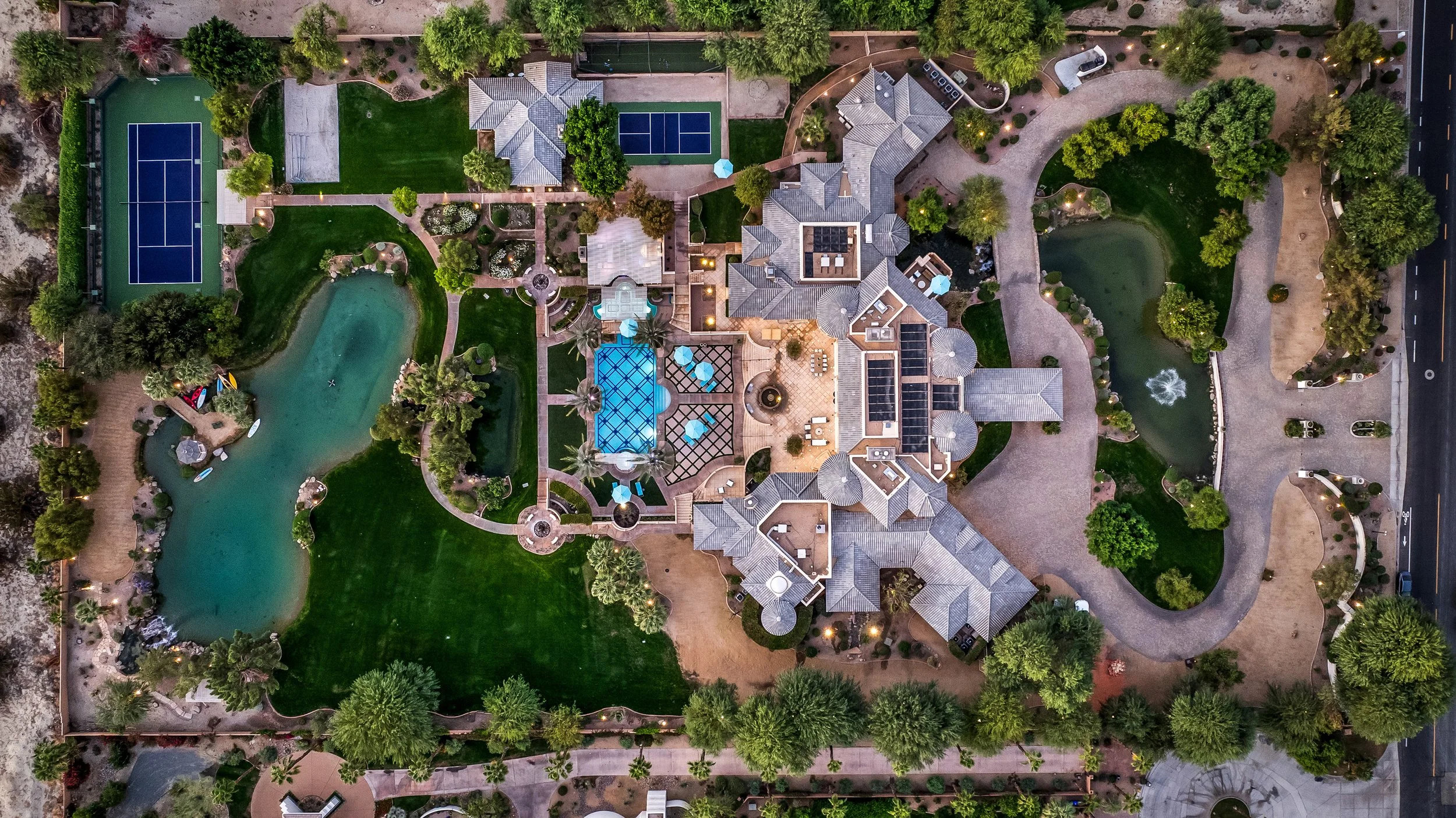 An aerial view of a large house with a swimming pool, multiple tennis courts, a pond with a fountain, and surrounding greenery and trees, with a driveway on the right side.