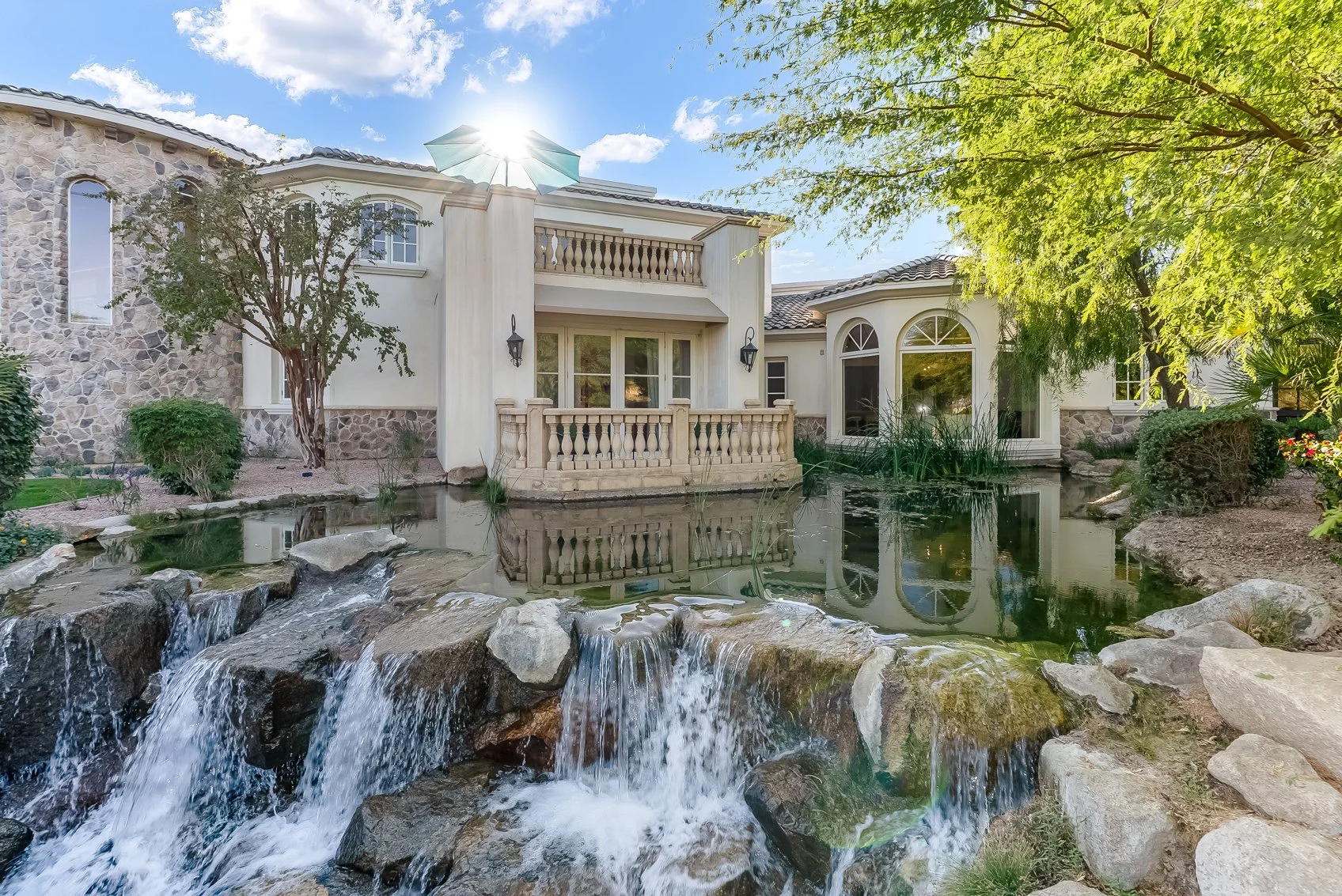 Luxury house with stone and stucco exterior, surrounded by lush trees and landscaping, beside a pond with a small waterfall in the foreground, under a bright blue sky with scattered clouds.