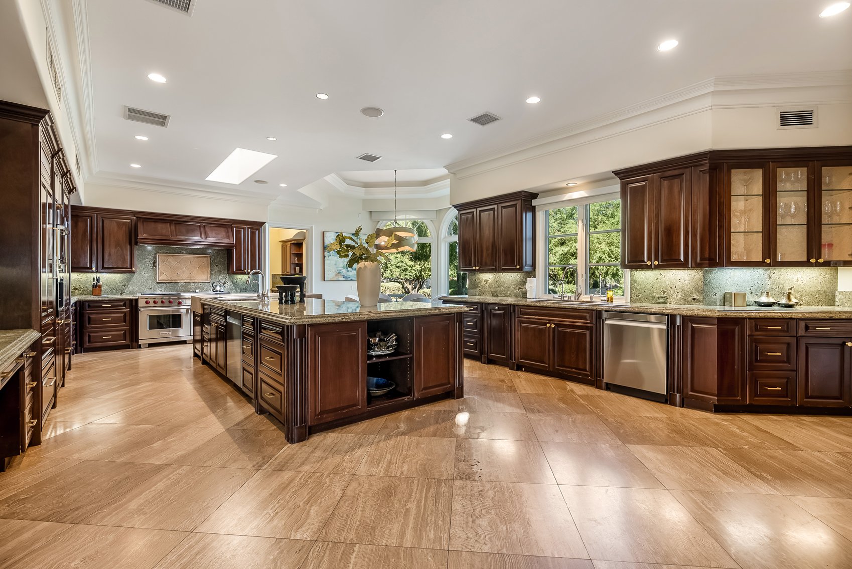 A spacious kitchen with dark wooden cabinets, a large island with a vase and decorative items, and stainless steel appliances, including an oven, dishwasher, and microwave. The room has a high ceiling with recessed lighting and large windows letting in natural light, with green trees visible outside.