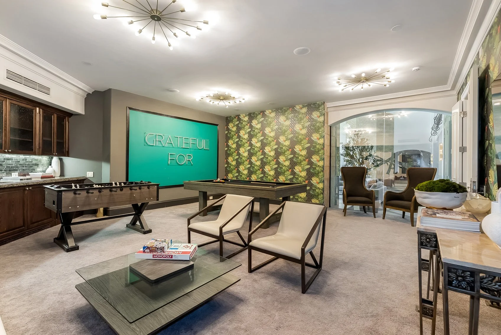 A modern recreational room with foam pool table, foosball table, seating area with two chairs, glass coffee table with Monopoly game, a large teal wall display with the words 'Grateful For', decorative wallpaper, and large windows with view of a patio.
