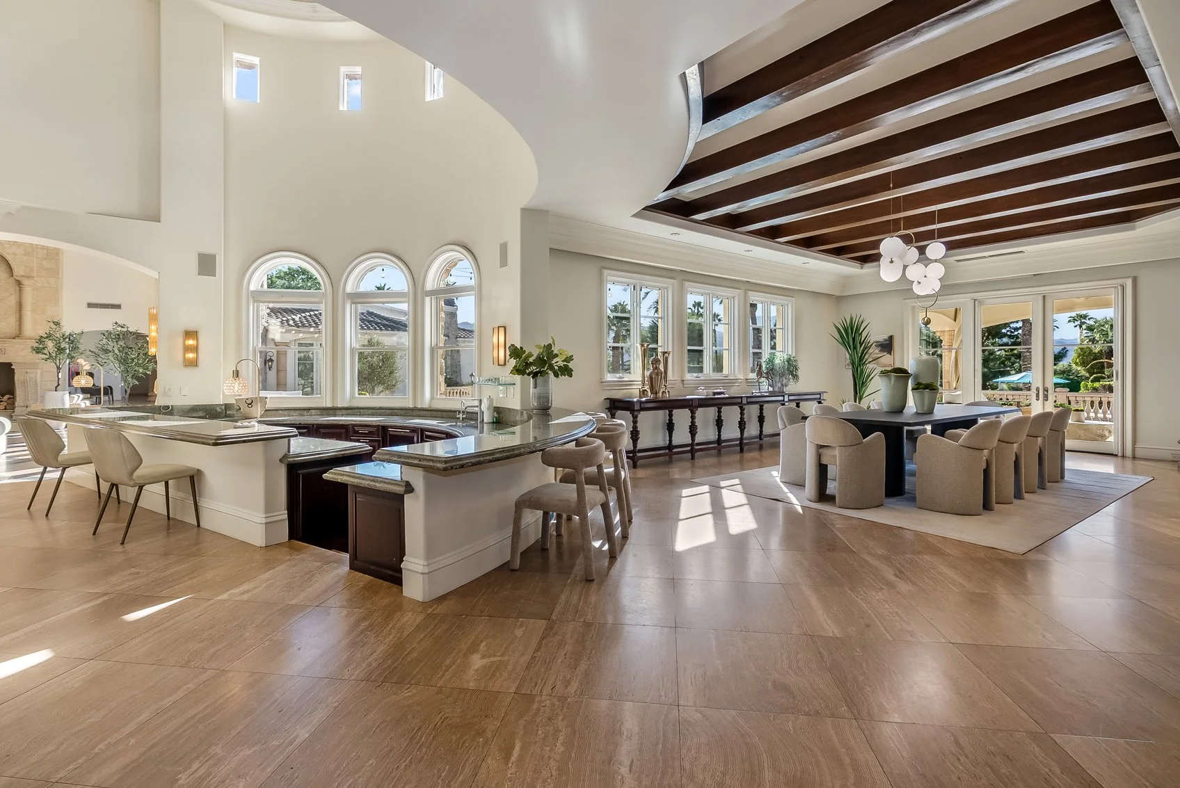 Spacious, sunny open-concept living and dining area with high vaulted ceiling, large windows, and hardwood floors. The room features a kitchen with a curved breakfast bar, a formal dining table with beige chairs, and a view of outdoor greenery through French doors.