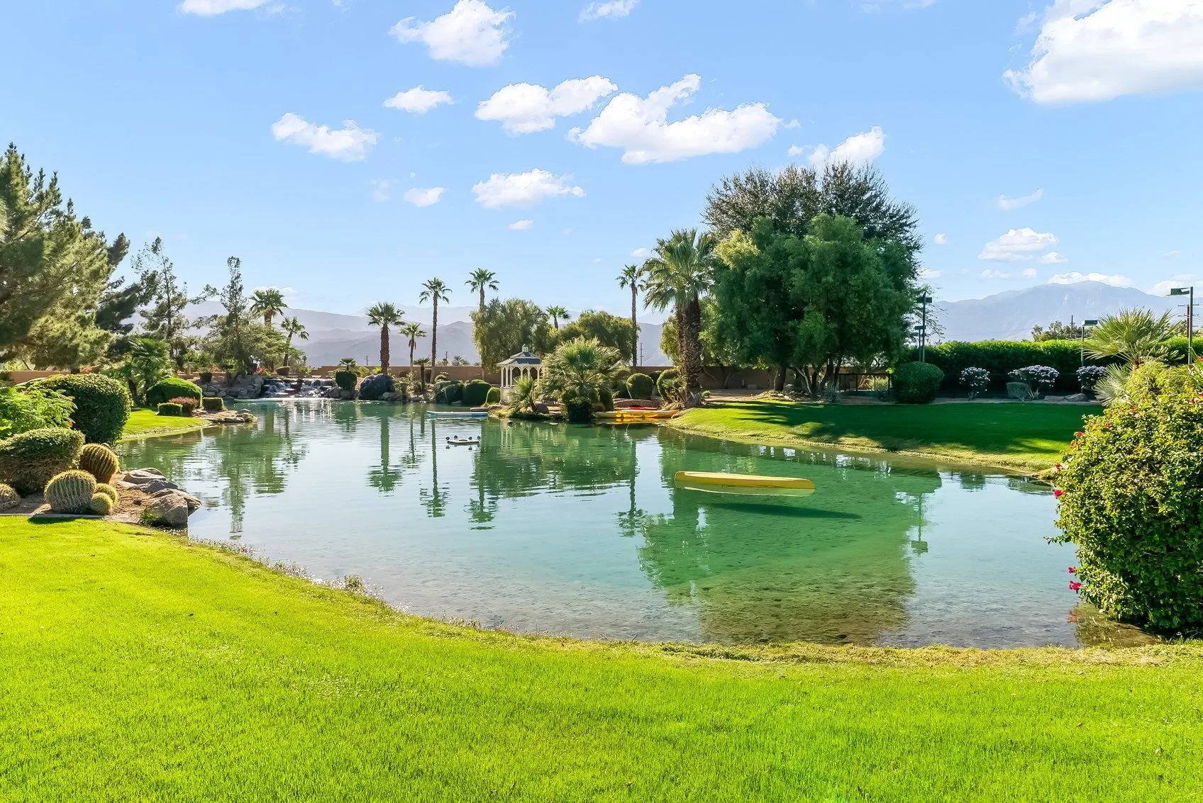 A tranquil park scene with a calm pond surrounded by green grass, various trees including palms, and desert plants like cacti. There are small boats on the water, a gazebo, and mountains in the background under a bright blue sky with scattered clouds.