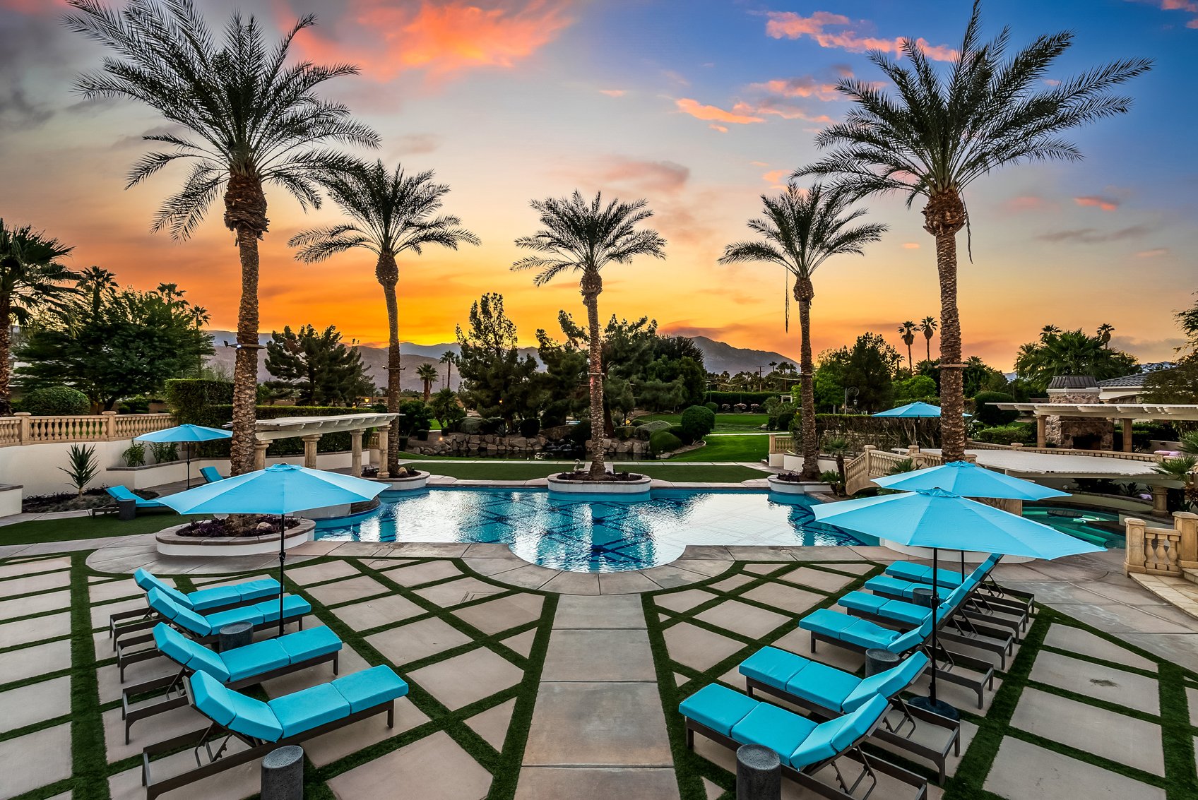 Luxury resort pool area during sunset with blue lounge chairs, matching umbrellas, palm trees, and mountains in the background.