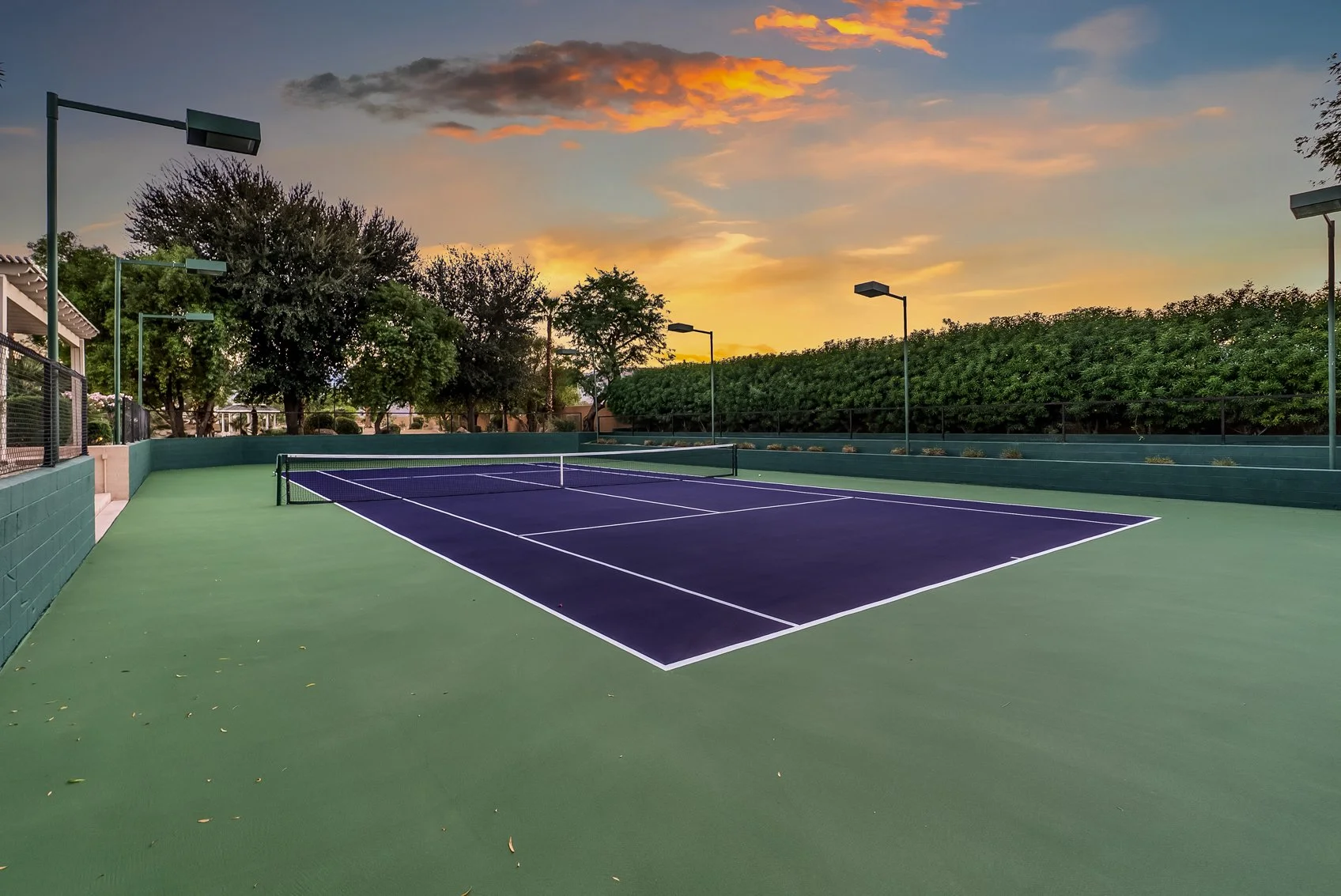 Empty outdoor tennis court at sunset, surrounded by trees and lampposts.