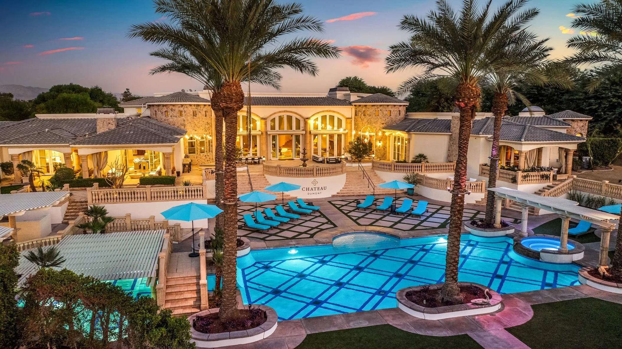 Luxury mansion with illuminated outdoor pool area surrounded by palm trees at sunset.