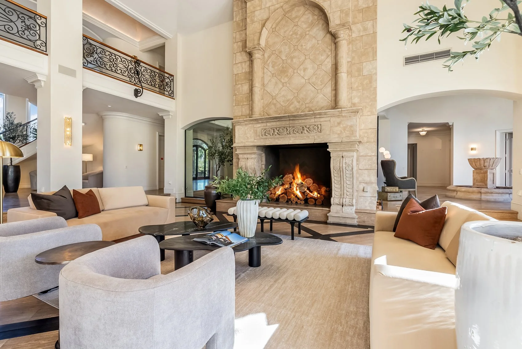 Elegant living room with a large stone fireplace, modern furniture, and a roaring fire. Bright and spacious with high ceilings, decorative railings, and a neutral color palette.