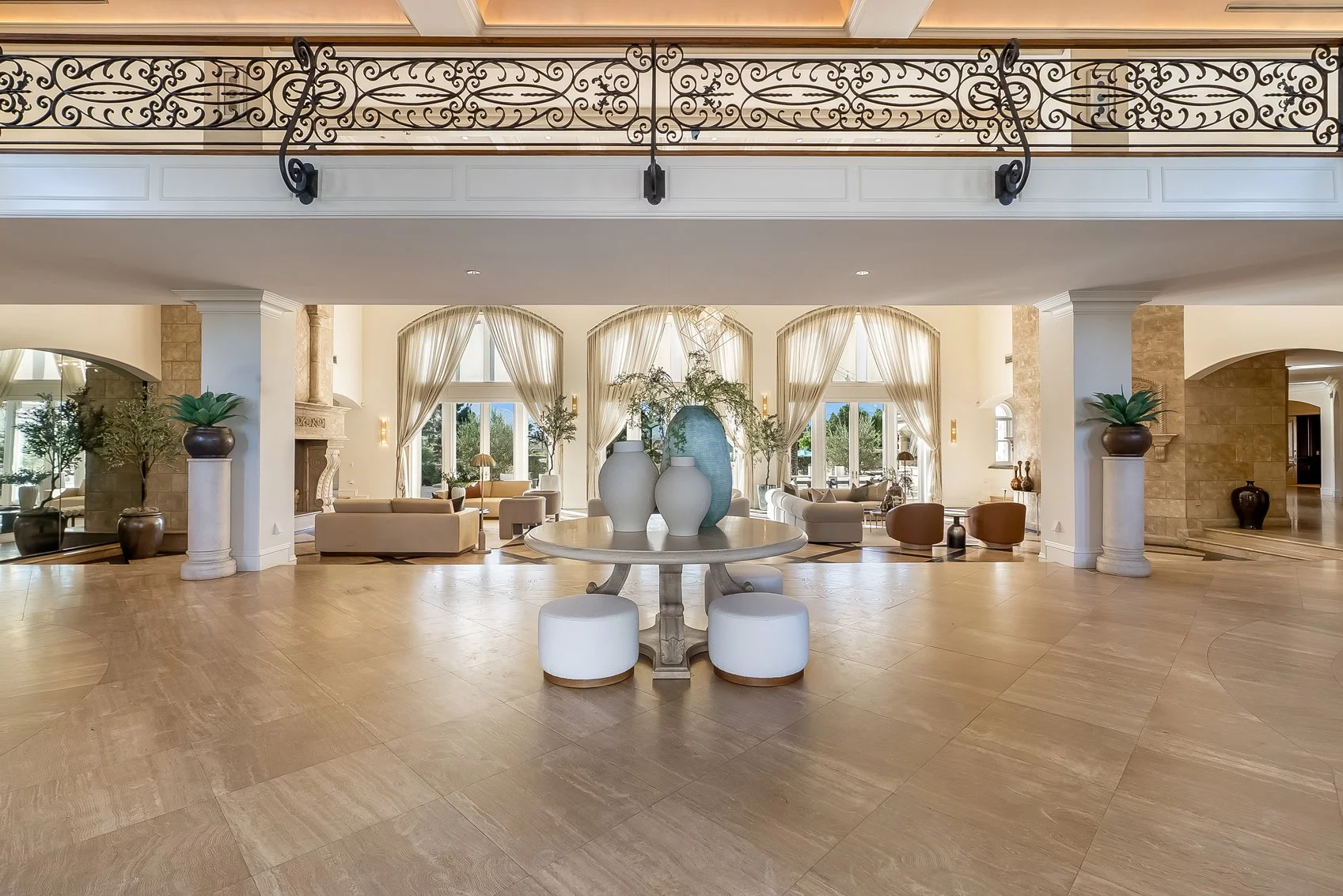 Spacious hotel lobby with high ceilings, large windows with draping curtains, and elegant furniture. A round table in the center holds decorative vases, and potted plants are on columns. Seating areas include sofas and armchairs with side tables, creating a bright, open atmosphere.