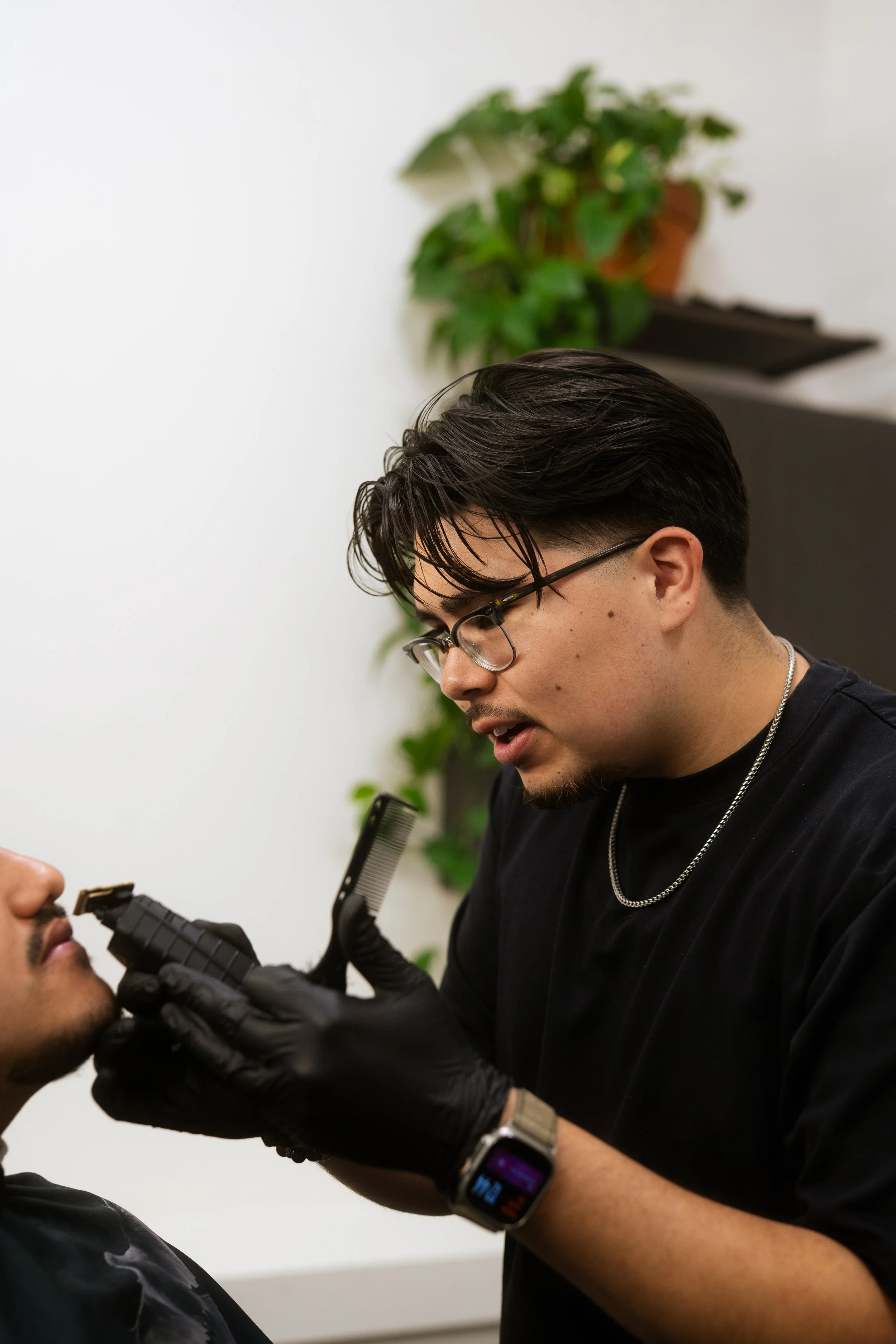 A man with glasses getting a close shave from a barber using a straight razor in a barbershop, with a plant and black furniture in the background.