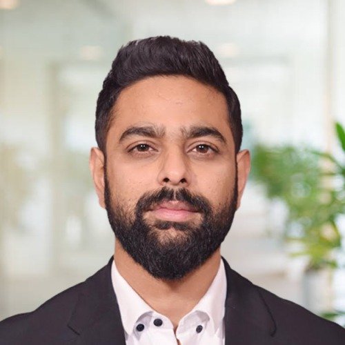A man with dark hair, beard, and mustache wearing a white shirt and black blazer, standing indoors with a blurred background that includes plants. Nakul Gandhi, lawyer, advocate, Karanjawala & Co.