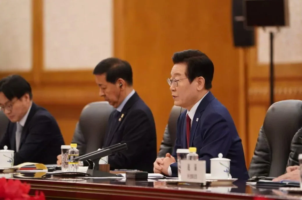 A group of men sitting at a conference table, with one man in a blue suit speaking, others with headsets, water bottles, and teacups in front of them.