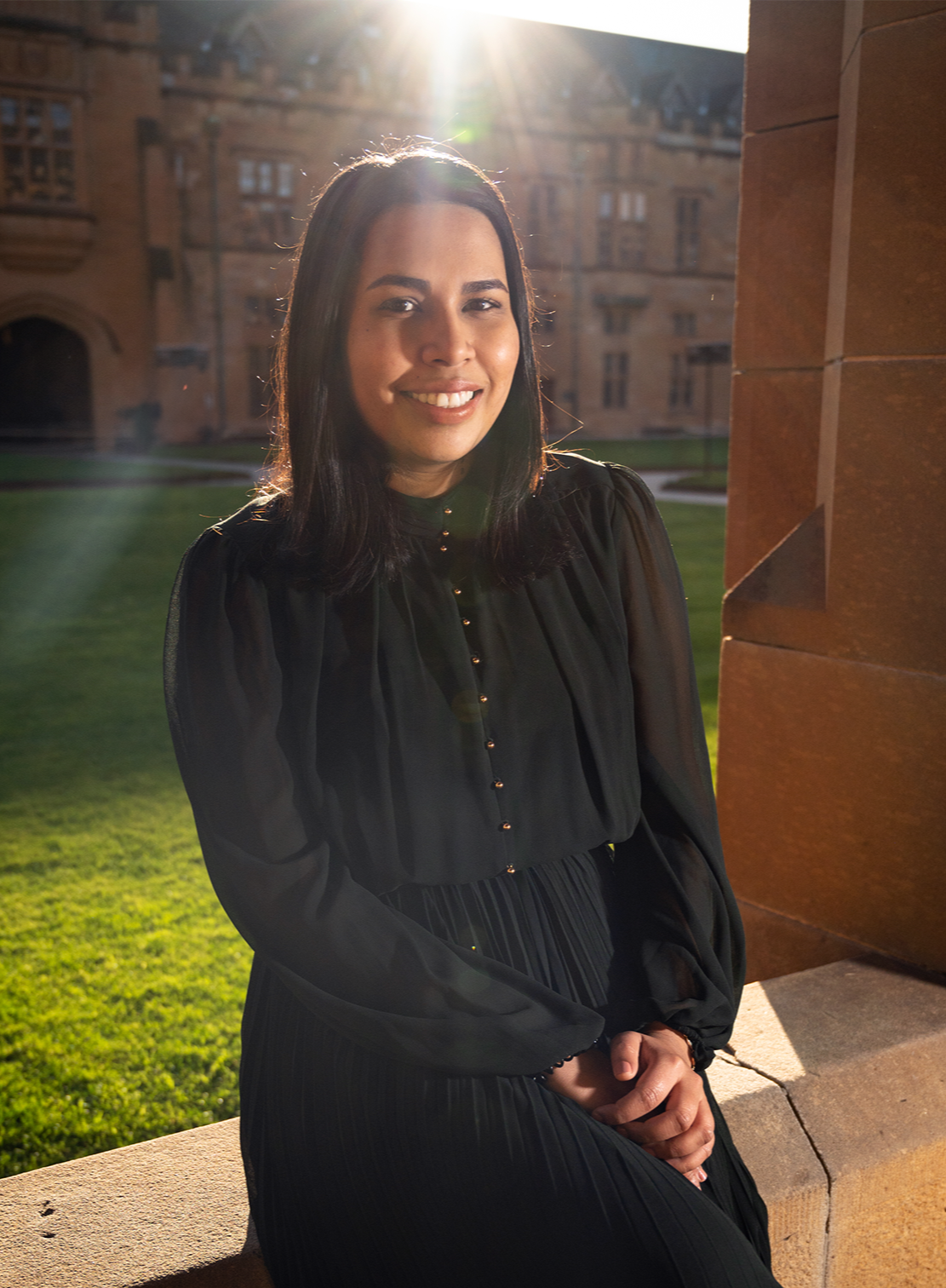 A woman with black hair and a black long-sleeve blouse sitting outdoors on a stone ledge with a historic brick building in the background, illuminated by sunlight.