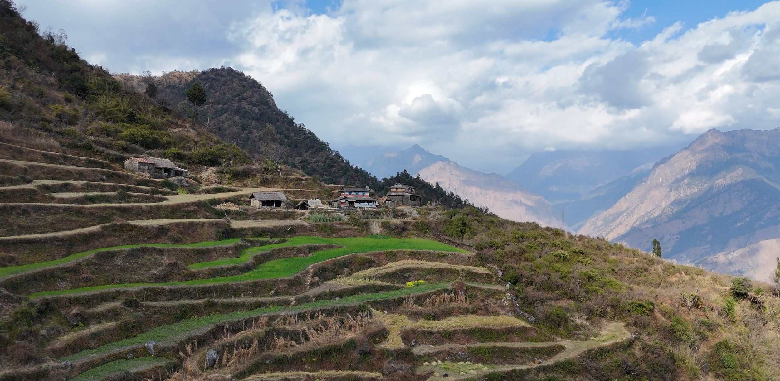 Terraced rice fields on a hillside with small houses and mountains in the background under a partly cloudy sky.