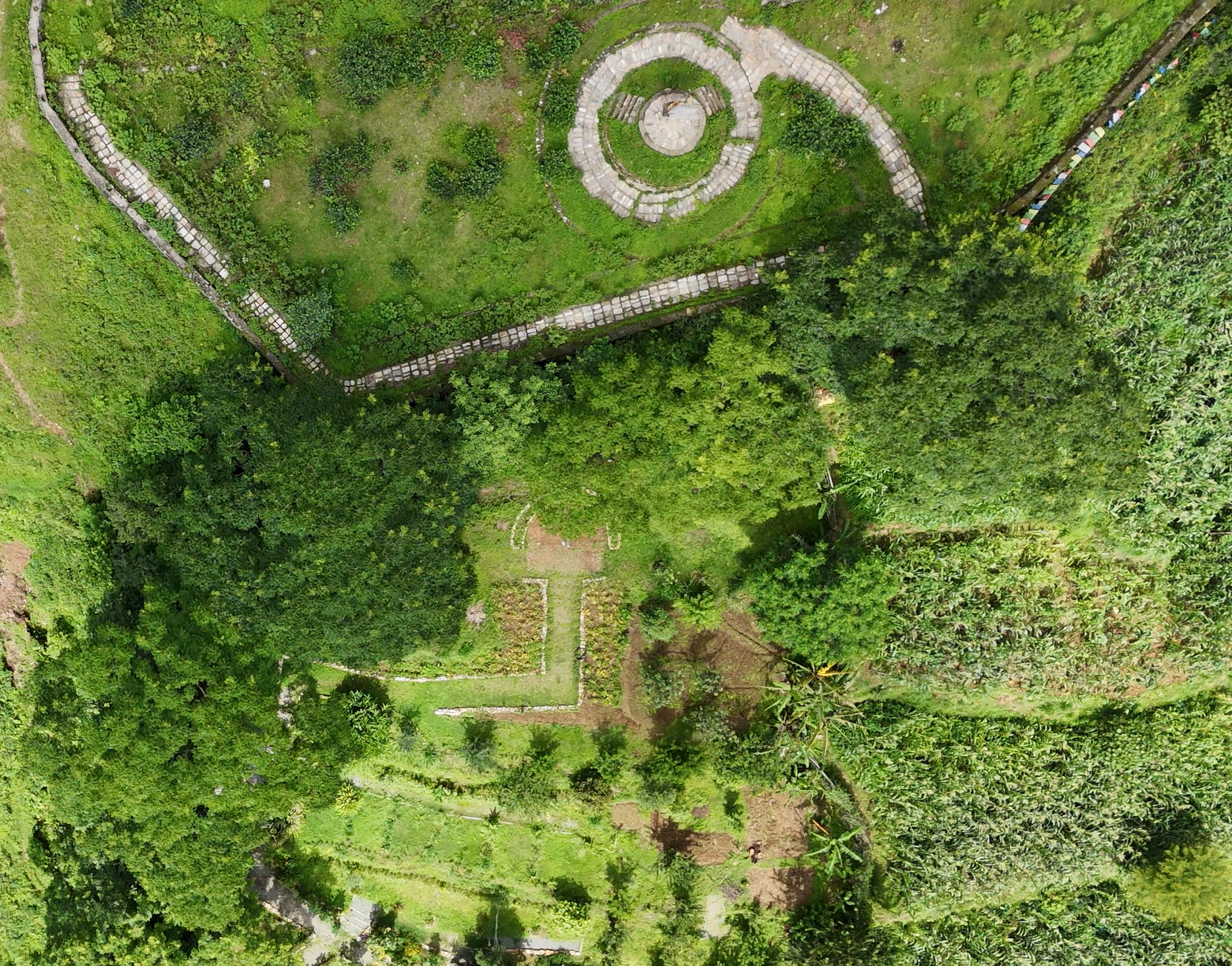 Aerial view of a lush green garden with winding pathways, a circular stone structure, and surrounding trees and vegetation.